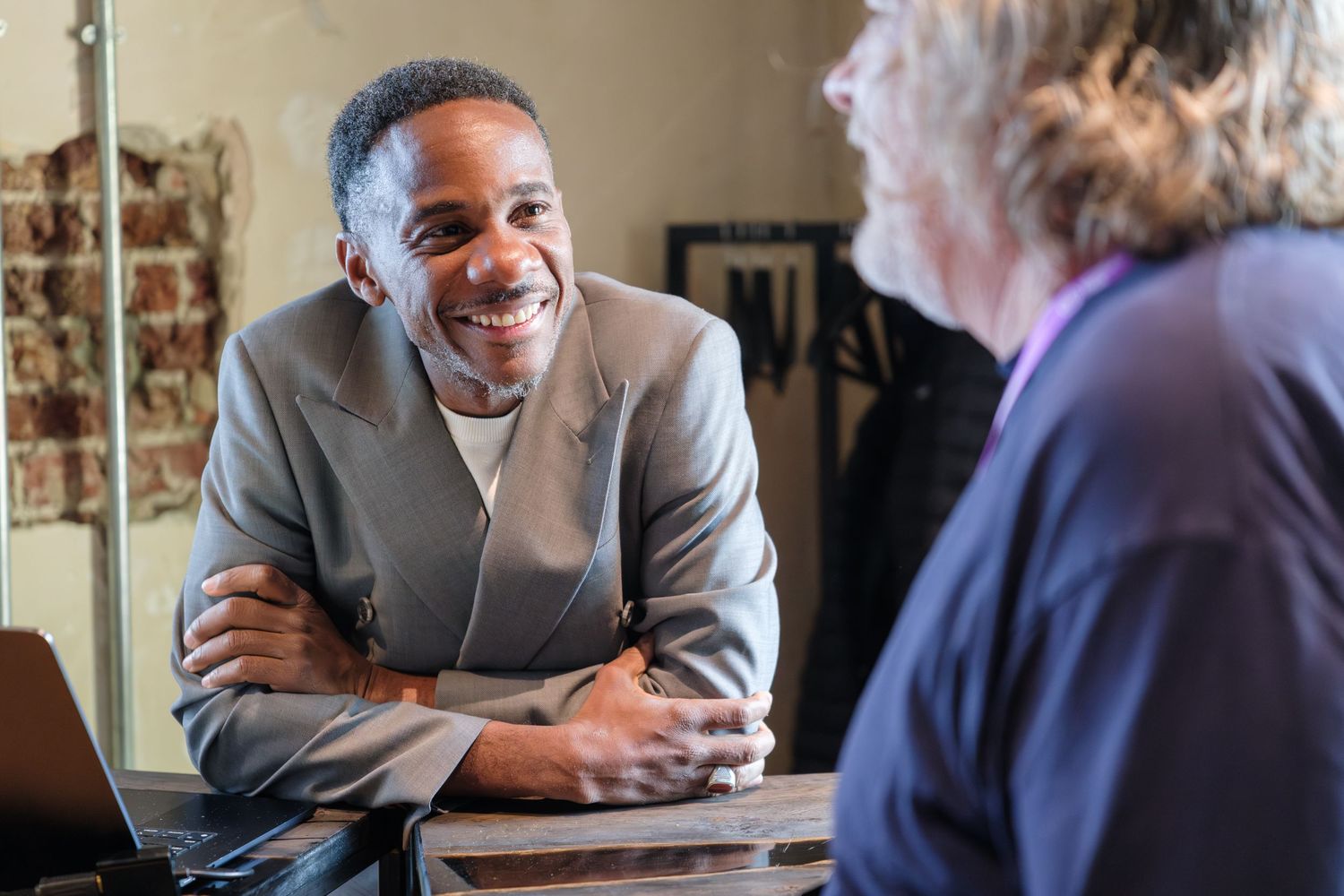 Two professionals engaged in a friendly conversation at a table, with a smiling man in a gray blazer listening attentively.