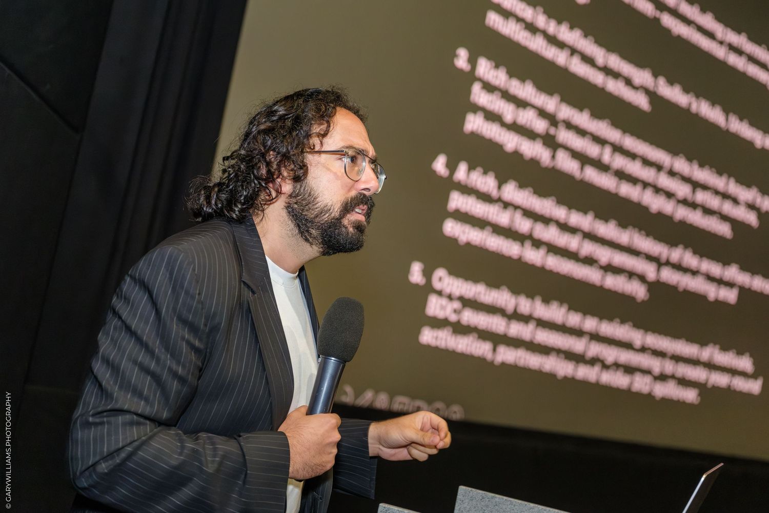 Bearded man in suit jacket speaking at conference podium with presentation screen behind him.