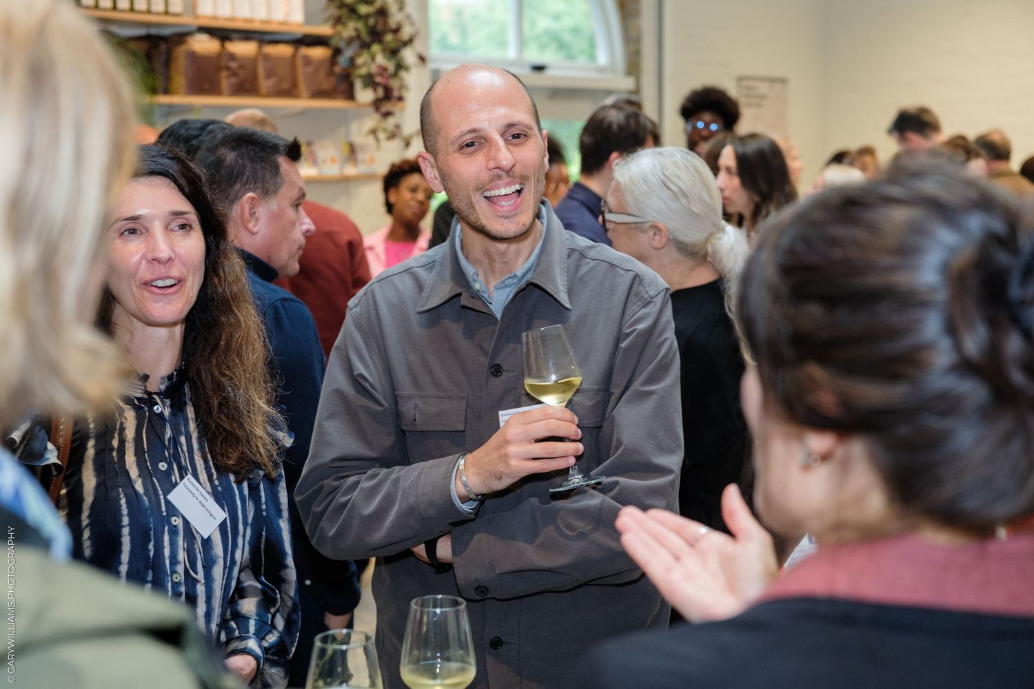 Man smiling and holding white wine glass at a social gathering event with guests mingling indoors.