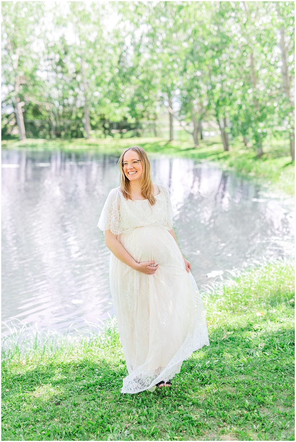 Expecting mother in flowing white lace dress poses gracefully by a serene pond surrounded by spring trees. Light & airy photography style.