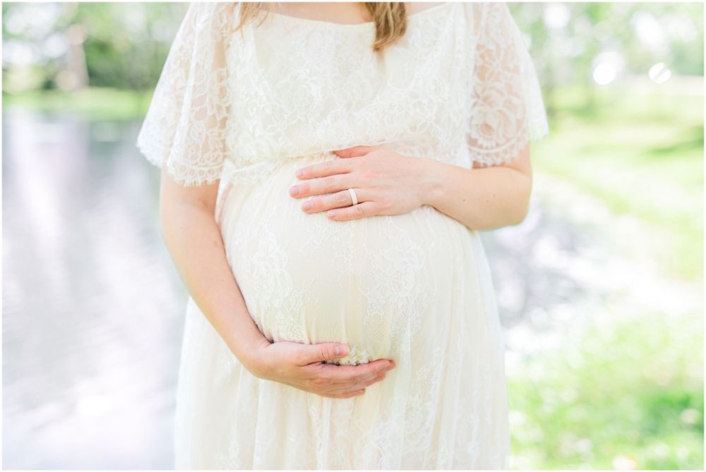 Close-up of hands cradling baby bump in flowing white lace maternity dress against natural backdrop. Light & airy photography style.