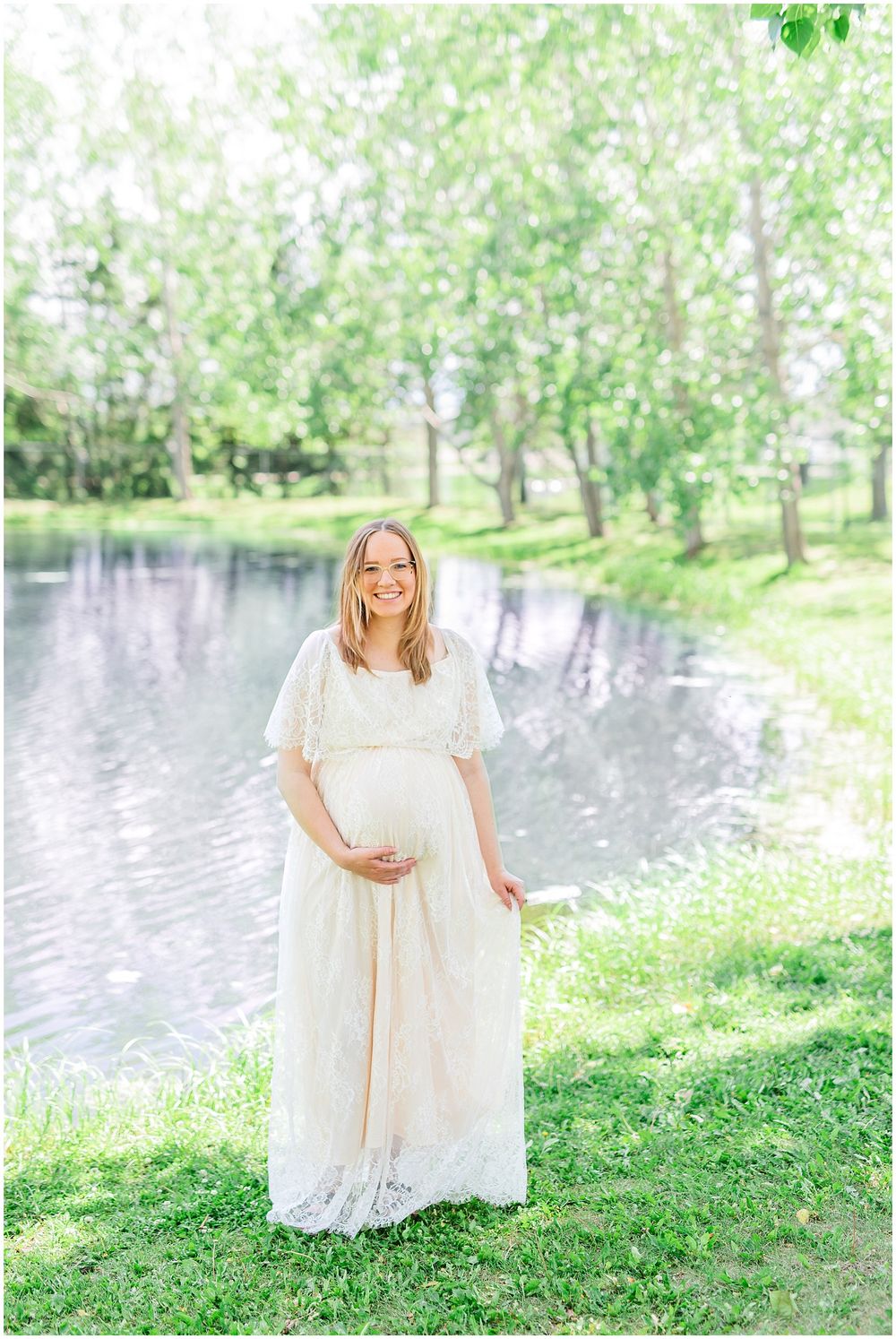 A maternity portrait in a flowing white lace dress against a serene pond backdrop.