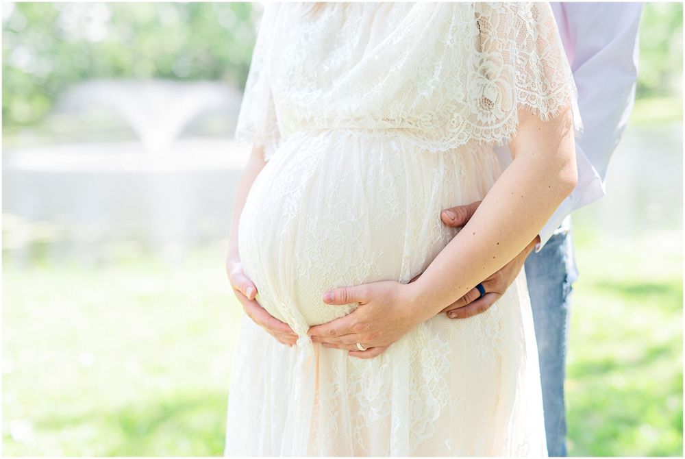 Close-up shot of hands cradling a baby bump in a delicate white lace maternity dress.