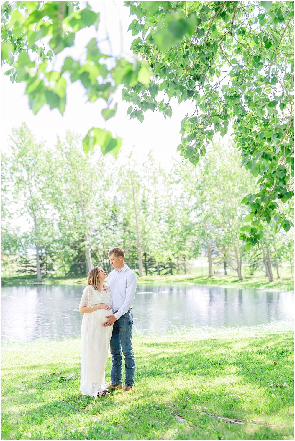 A romantic spring scene with a couple standing by a pond surrounded by lush green trees. Light & airy photography style.