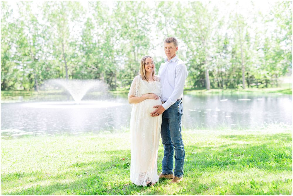 A serene maternity portrait by a fountain pond with fresh spring foliage in the background. Light & airy photography style.