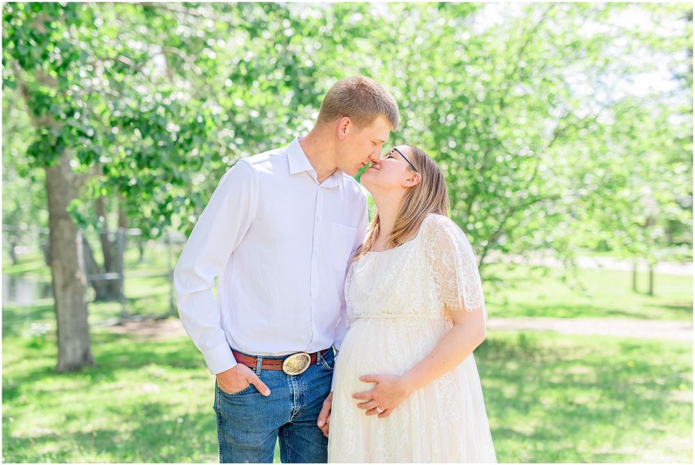 Intimate moment between expecting couple in white and casual attire surrounded by green spring foliage.
