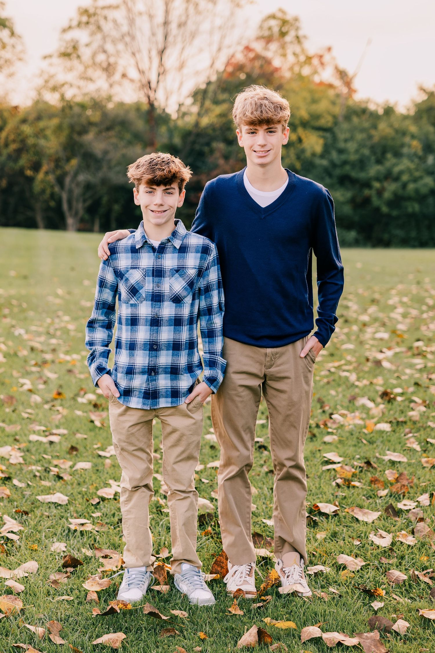 Two brothers in coordinating blue tops pose together on autumn grass with fallen leaves in Livonia Michigan.