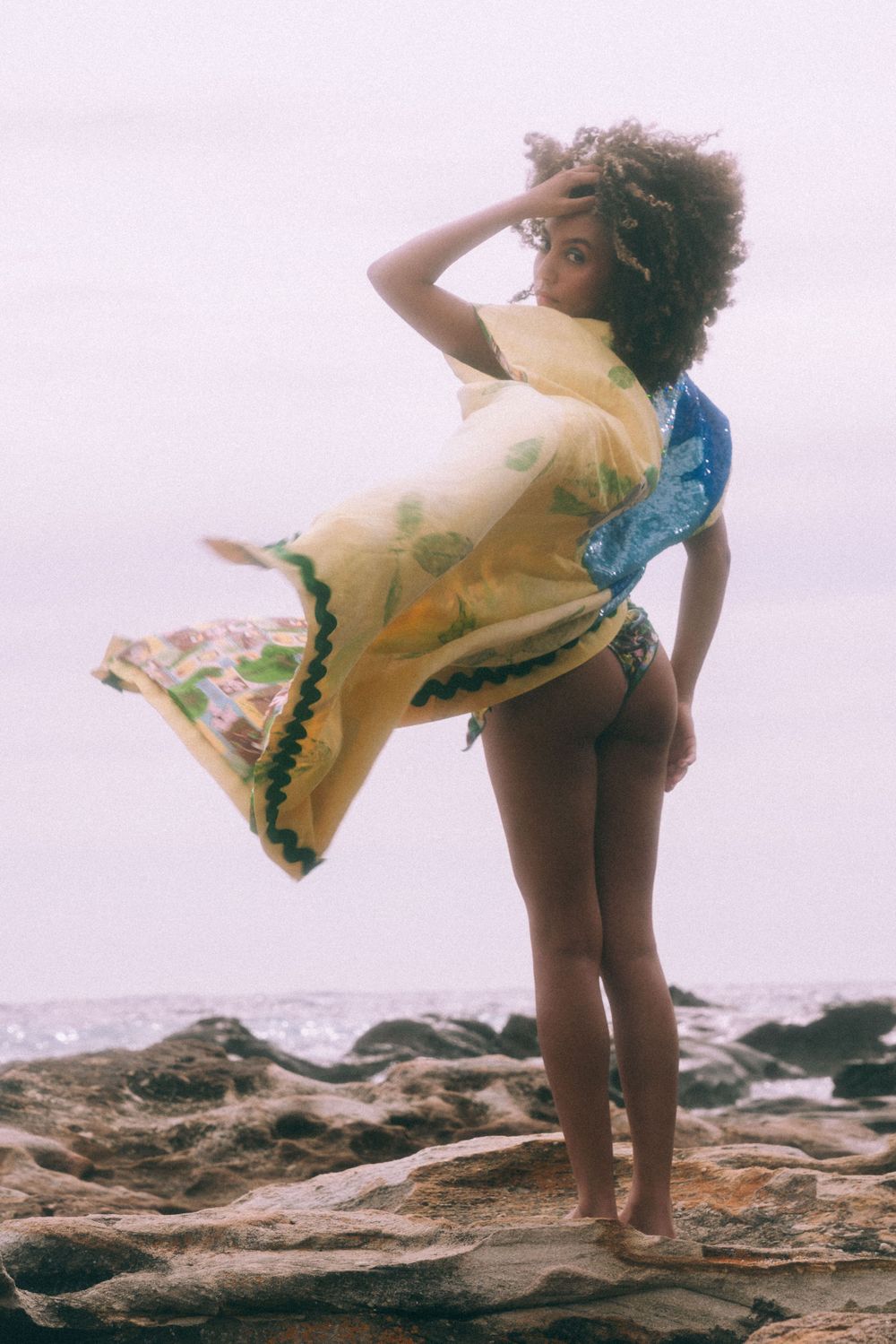 Fashion model in flowing dress poses on rocky beach overlook.