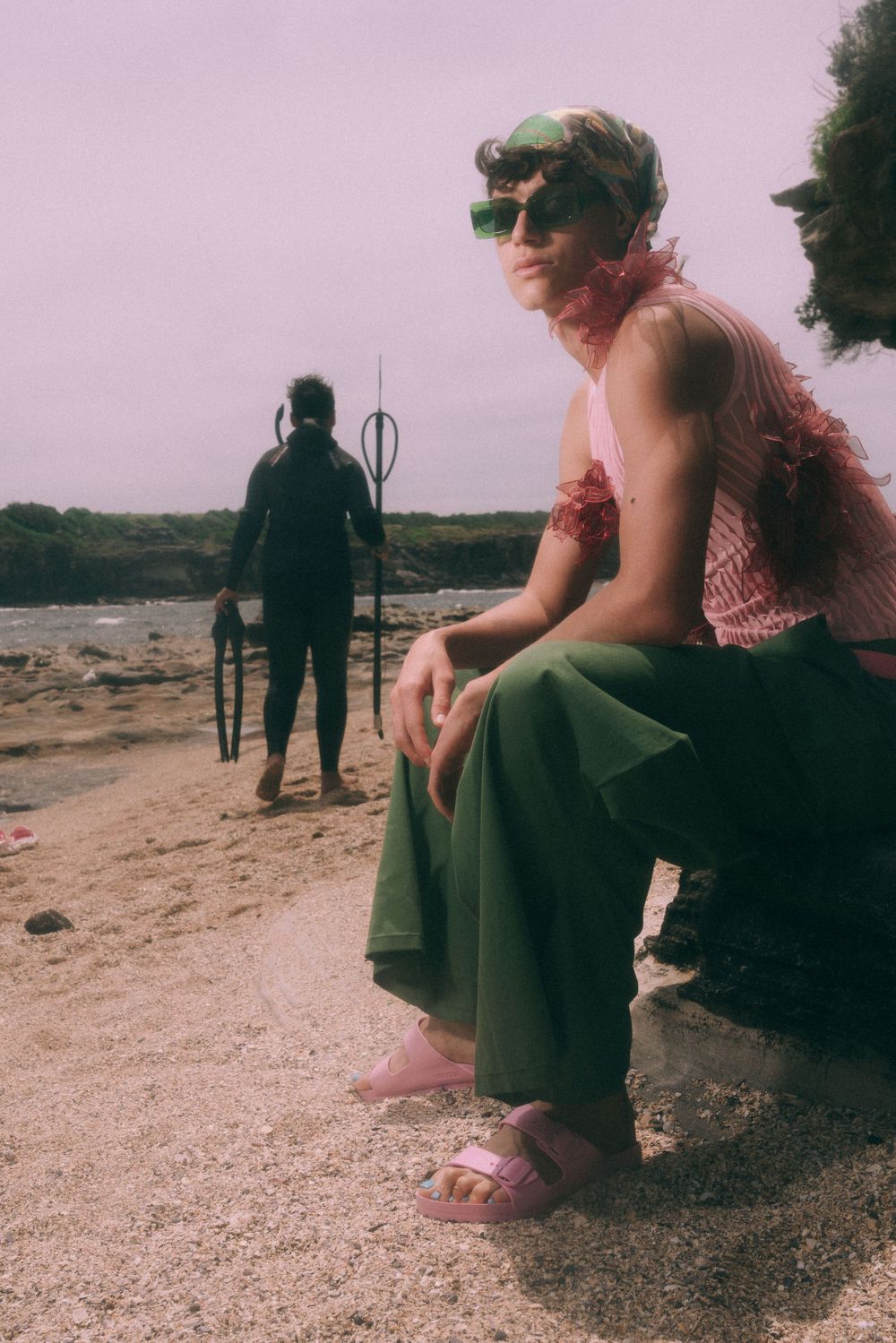Person in vintage-style outfit sitting on rocky terrain with distant figure in background against pink sky.