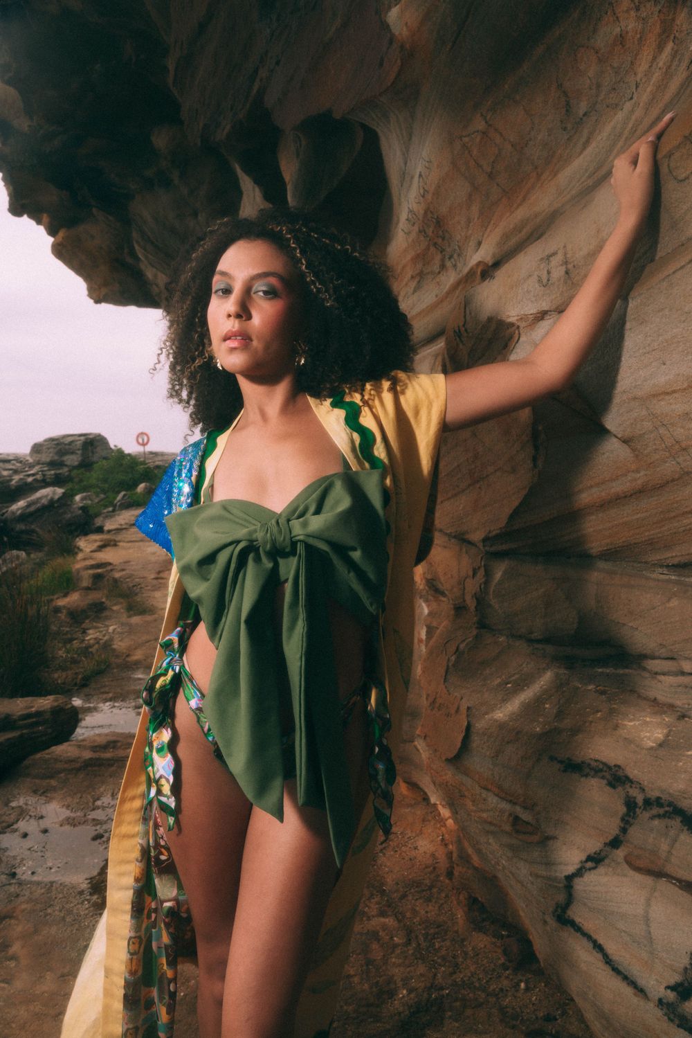 Model in green wrap poses dramatically against textured rock formation at sunset.