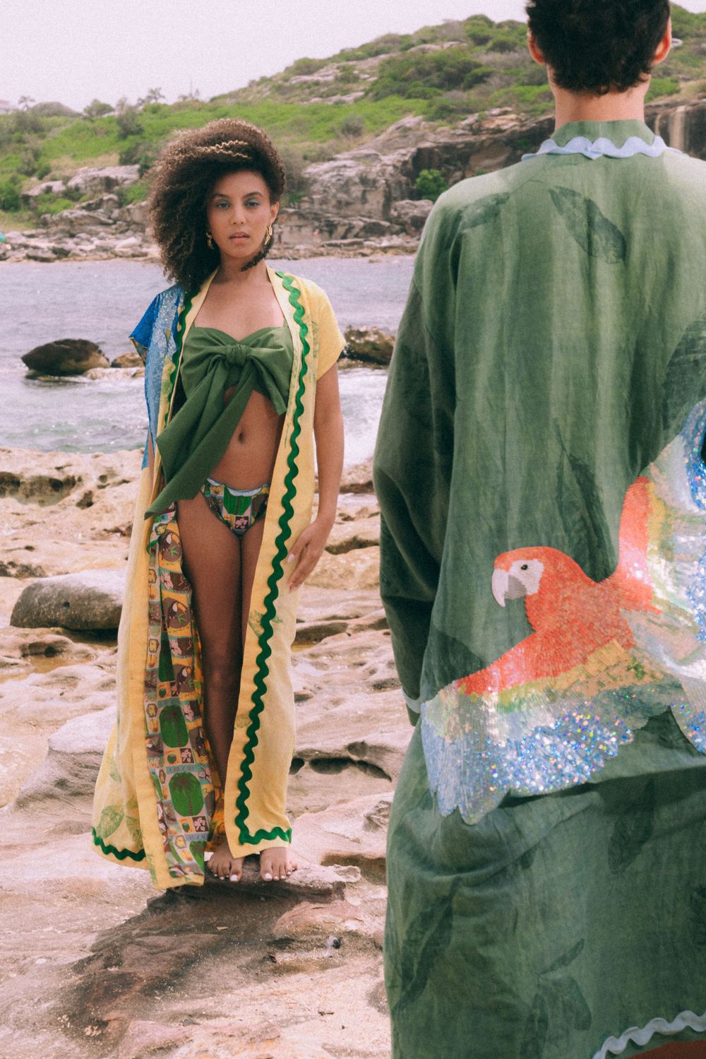 Two people in beachwear standing on rocky shore, one wearing parrot-printed robe.