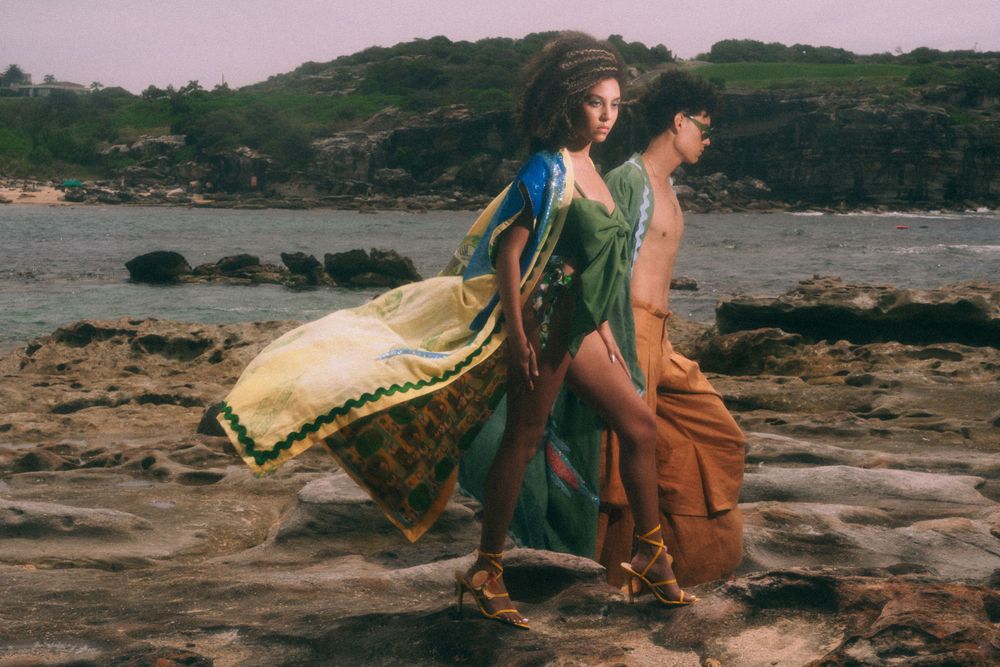 Models in flowing beach attire pose against rocky coastal landscape.
