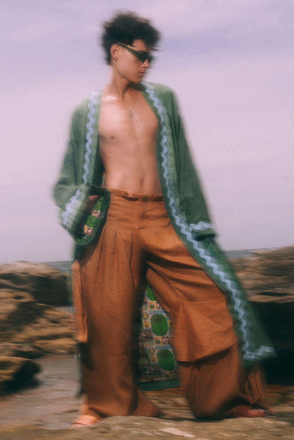 Model wearing green patterned jacket and wide-leg pants on rocky terrain.