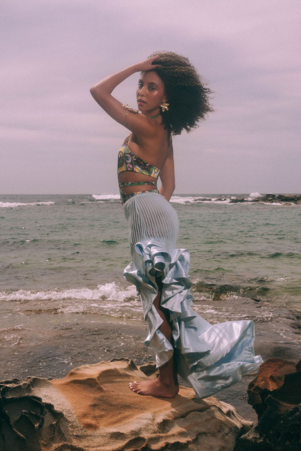 Beachwear fashion shot of model in metallic skirt posing on coastal rocks at sunset.