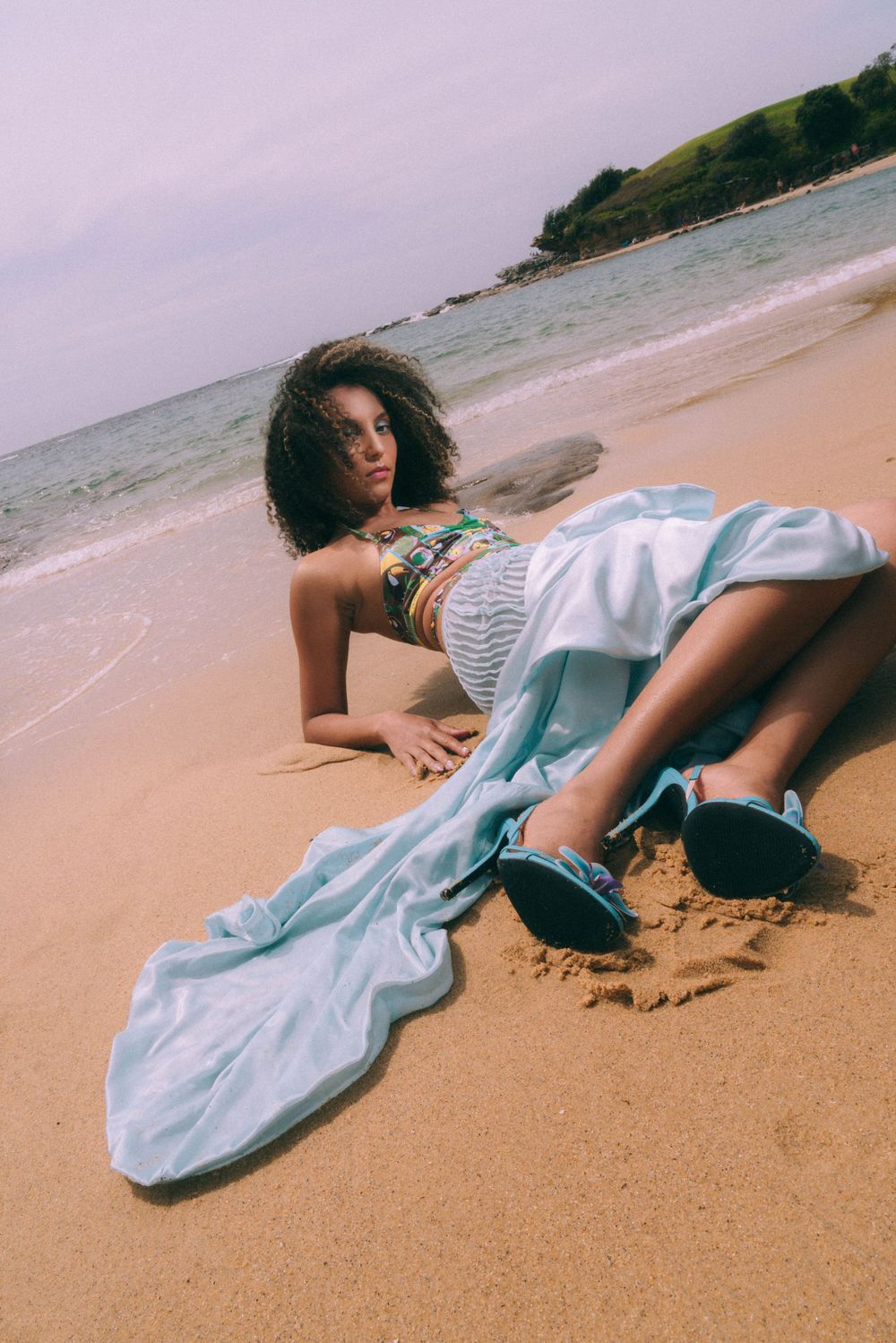 Beach fashion photo shoot with model reclining on sandy shore in summer attire.