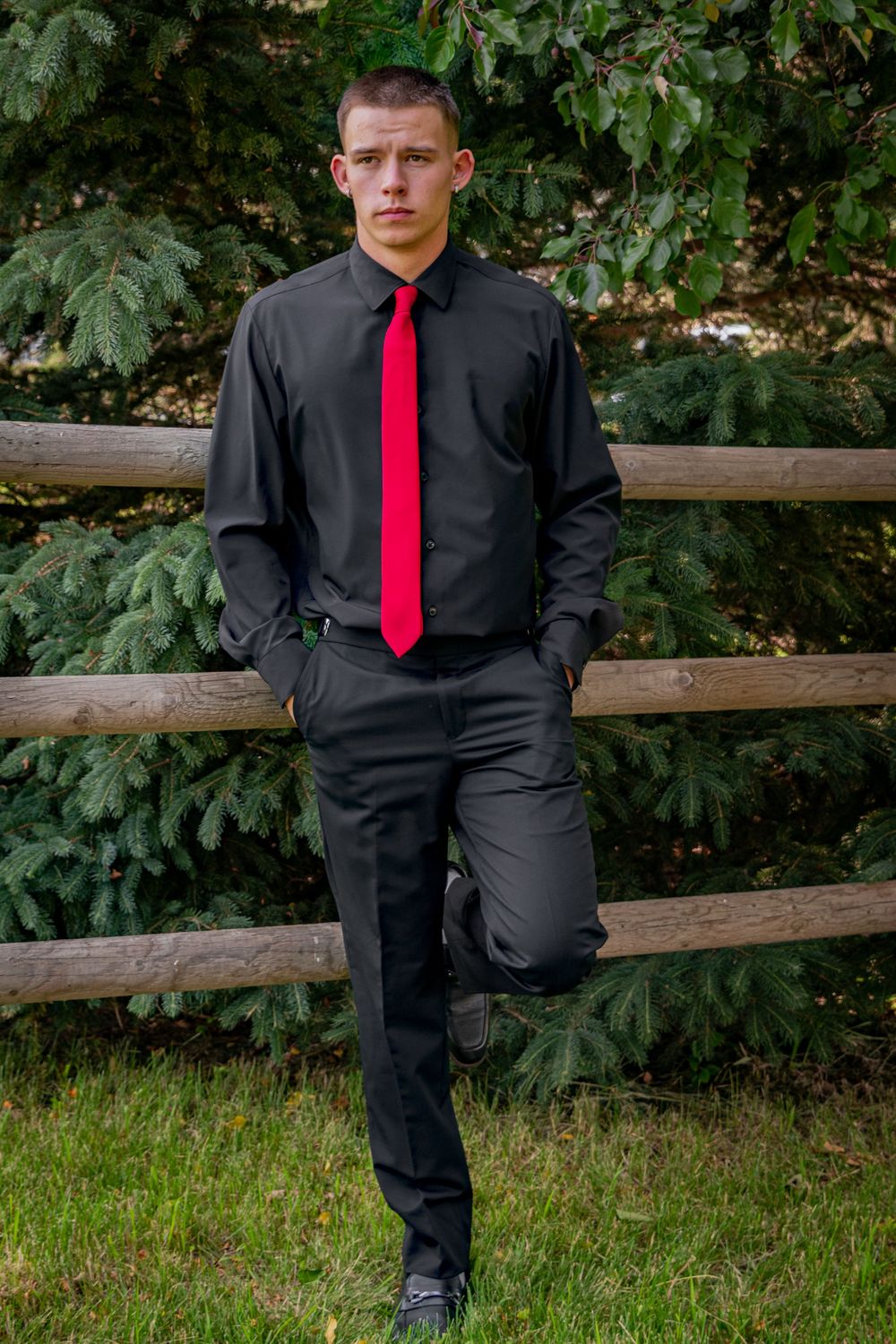 a young man in a tux leans on a wooden fence.