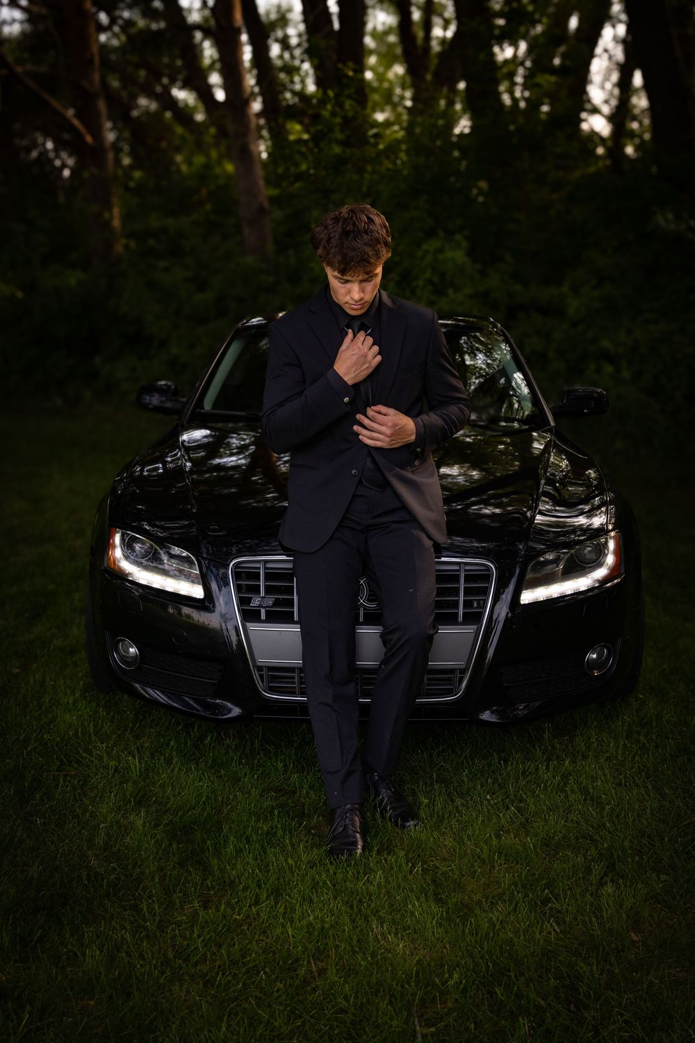 a young man in a tux leans on the hood of his car.
