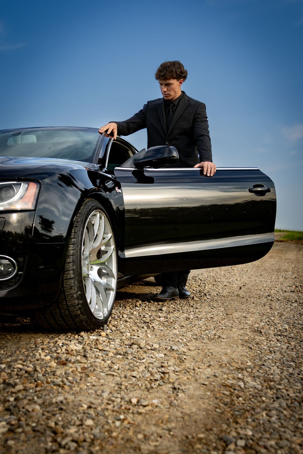 a young man in a tux stands with his car on a dirt road.