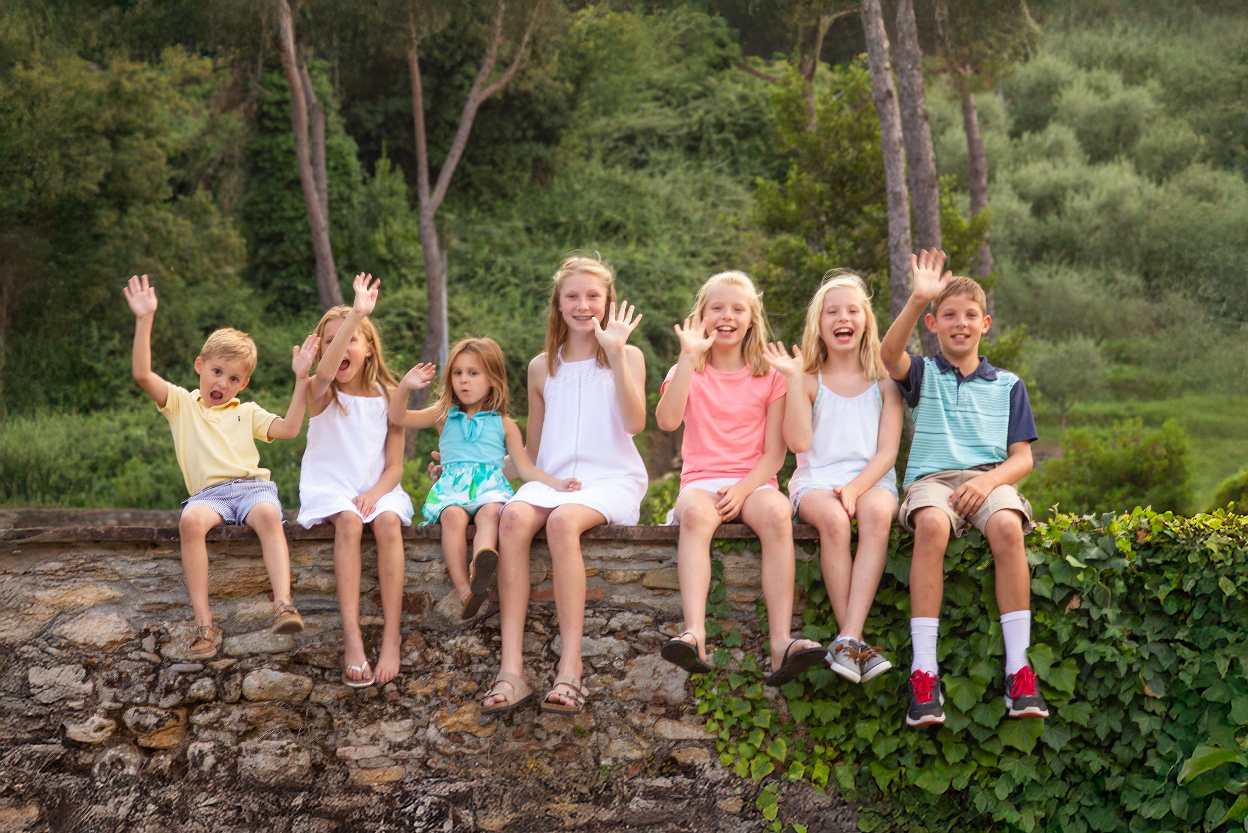 A group of children sitting together on a stone wall in a garden setting with greenery in the background.