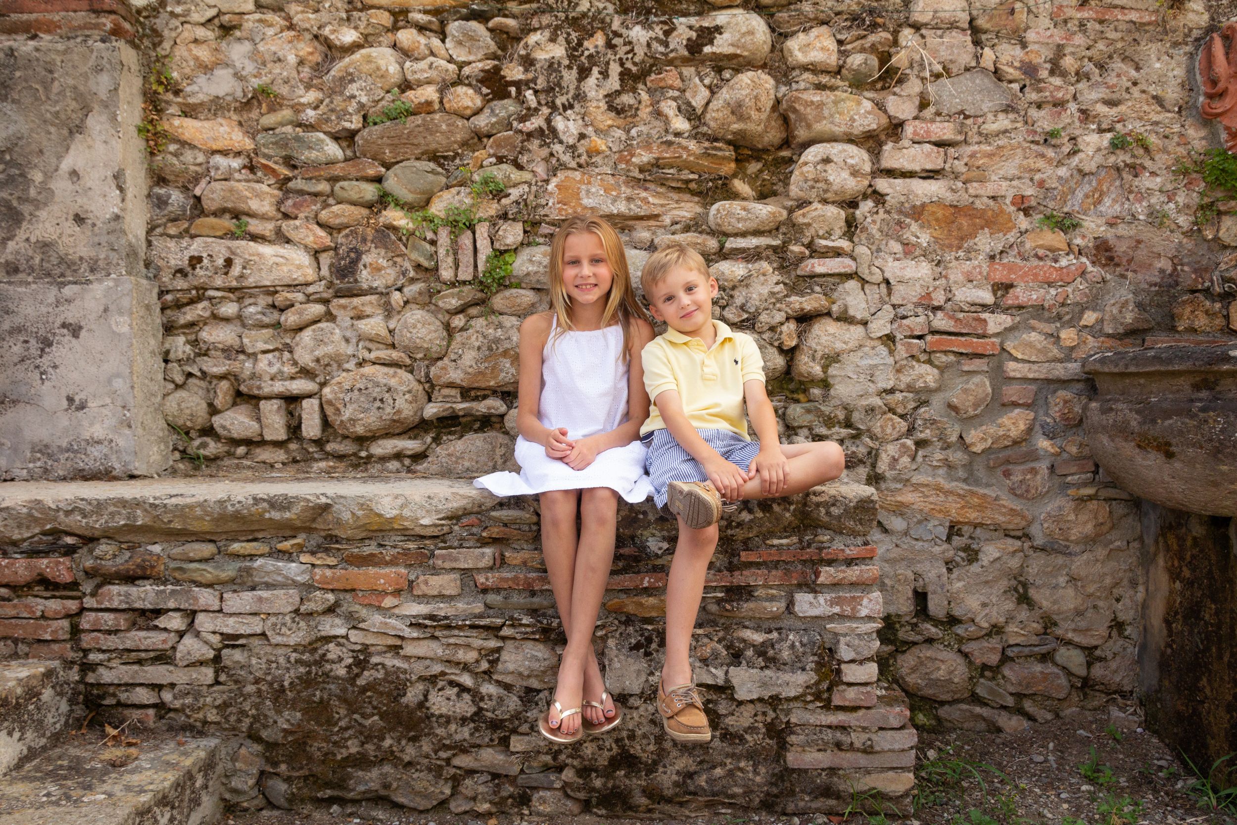 Two children sitting together on a rustic stone ledge against an old weathered stone wall outside.