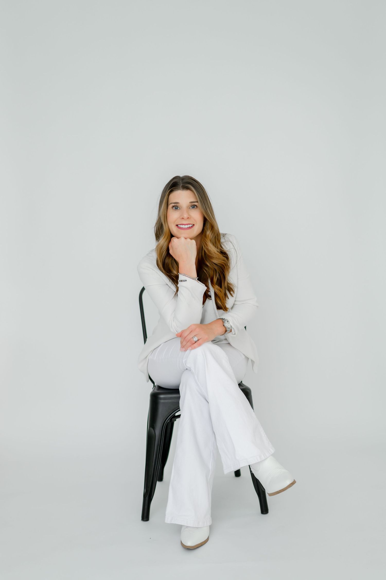 A person wearing all white clothing sits casually on a black stool against a light gray backdrop.