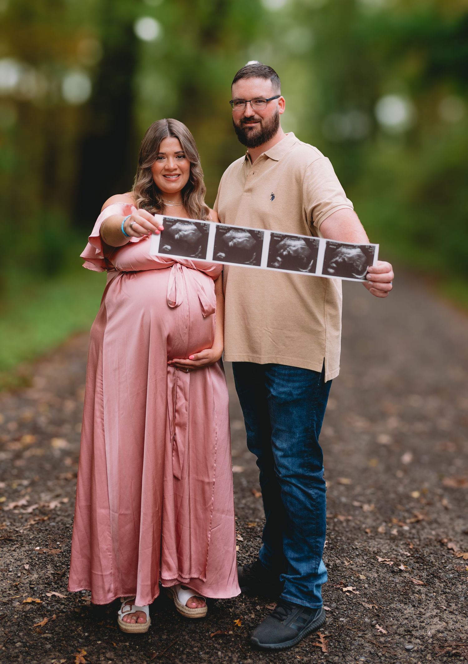 Couple holding ultrasound photos on a wooded path, with person in pink maxi dress showing pregnancy bump.