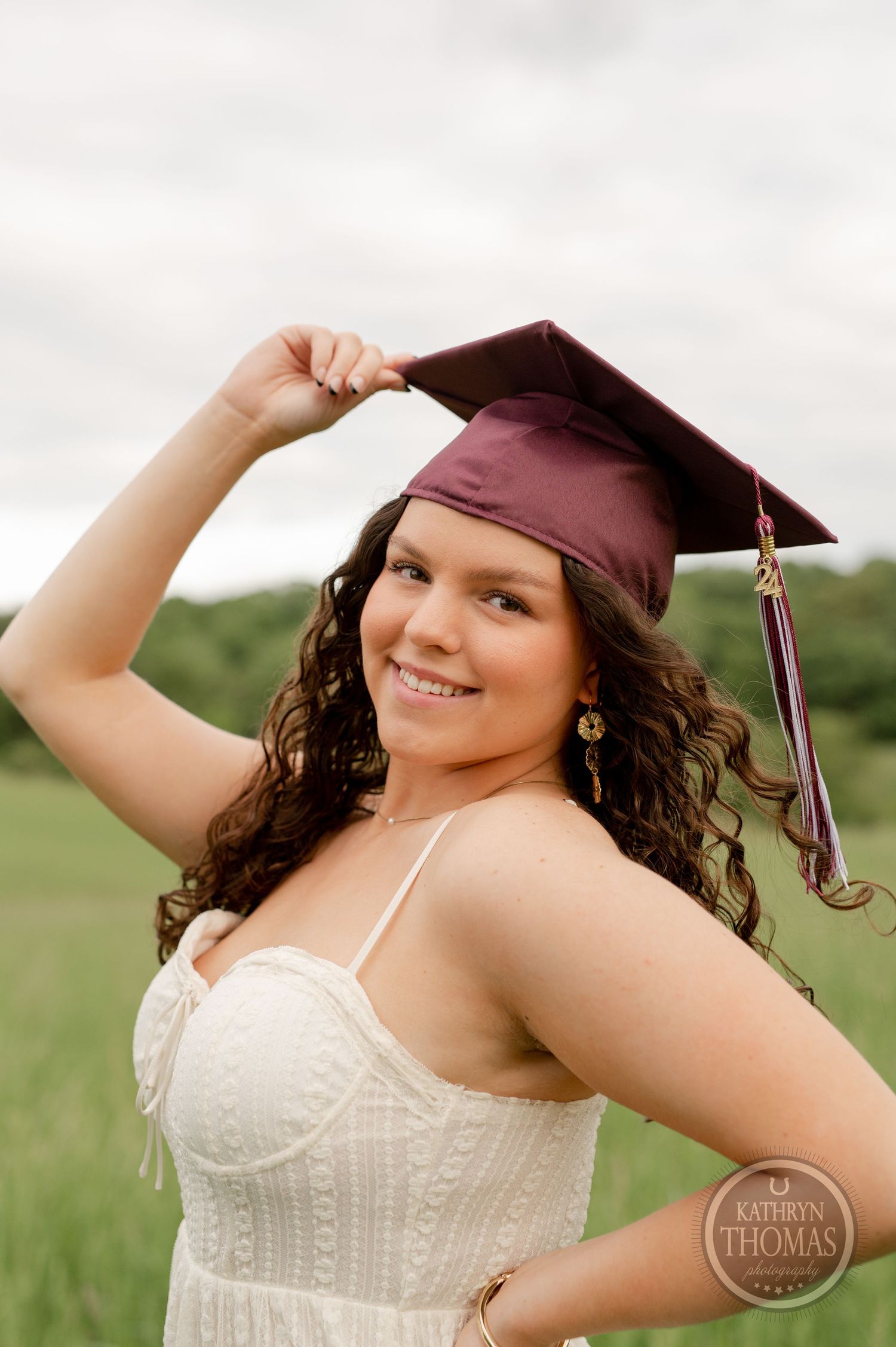 Graduate touches maroon cap while smiling in white dress outdoors.