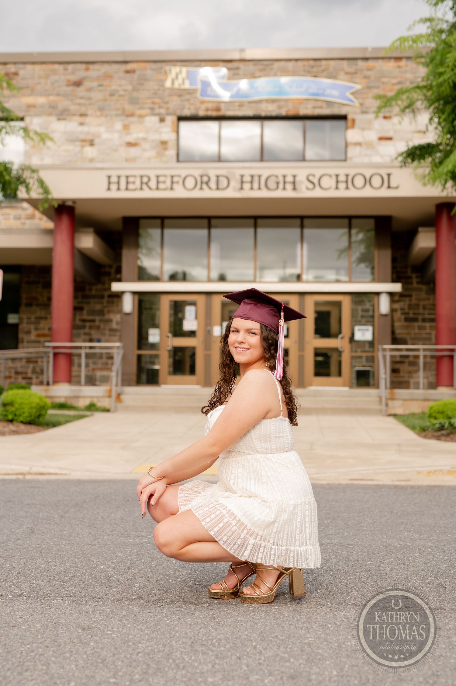 Graduate poses outside Hereford High School entrance in burgundy cap and white dress.