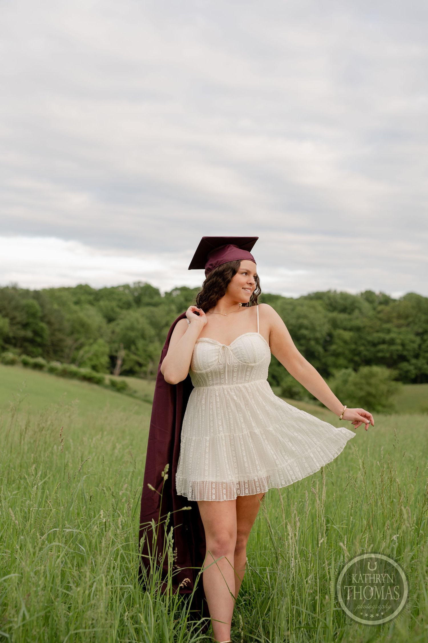 Graduate in a white dress twirls in a grassy field while wearing a burgundy cap and gown.