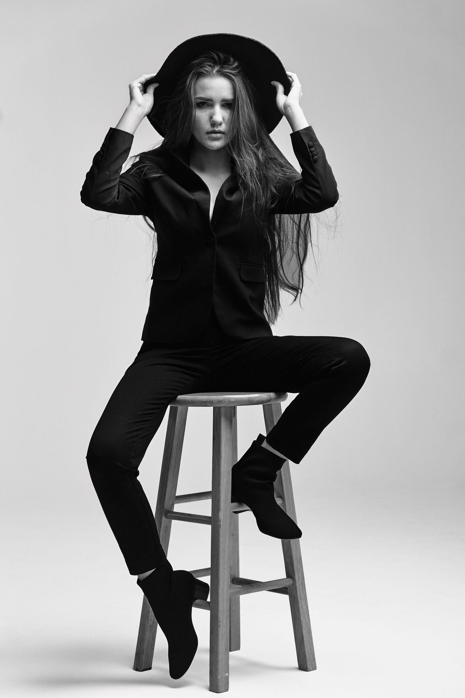 Artistic black and white portrait of figure in black outfit and hat seated on wooden stool.