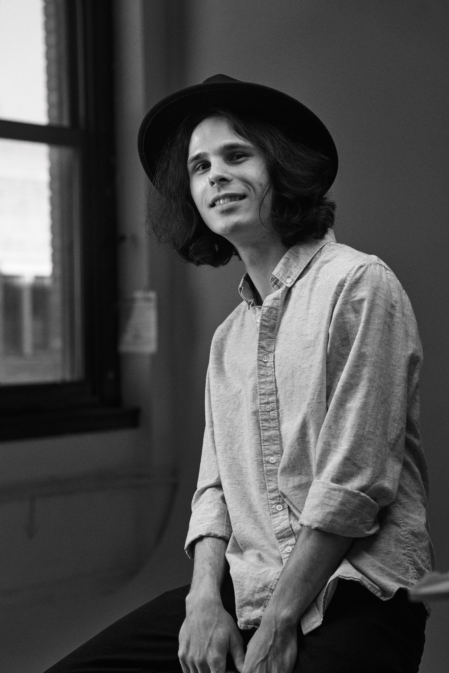 Black and white portrait of person in casual attire and wide-brimmed hat by window.