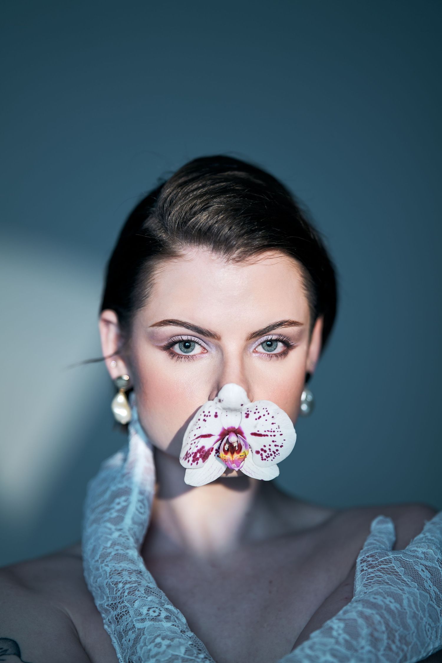 Artistic portrait with delicate orchid flower covering mouth against blue backdrop.