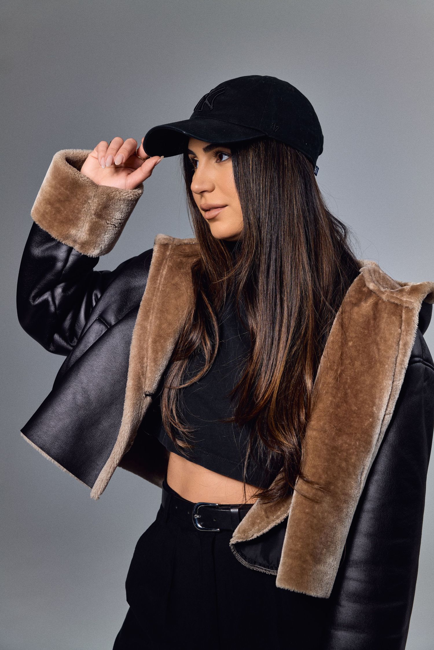 Model in black cap and fur-trimmed jacket poses on grey background.