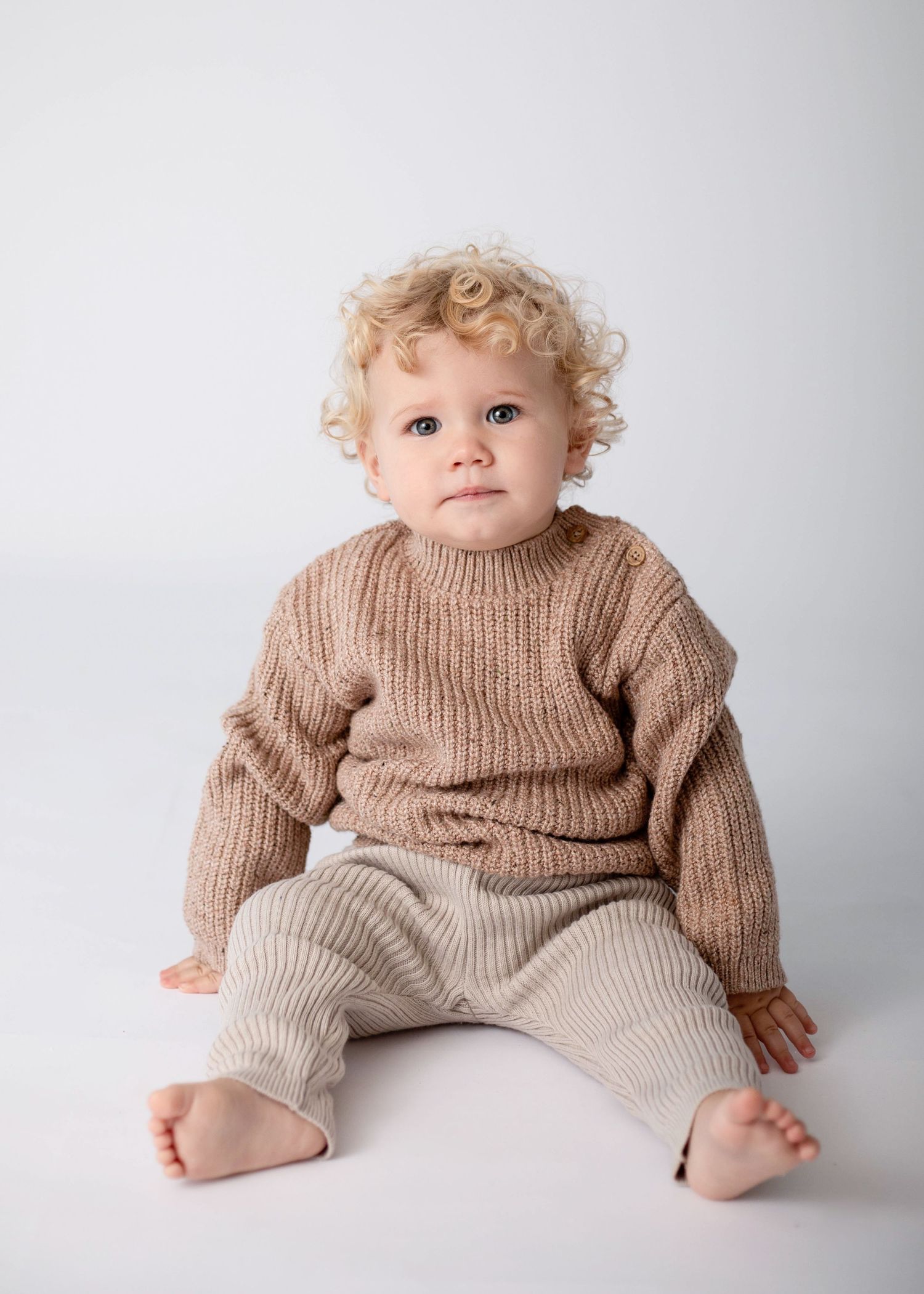 Young child wearing brown sweater and beige pants sits against white background.