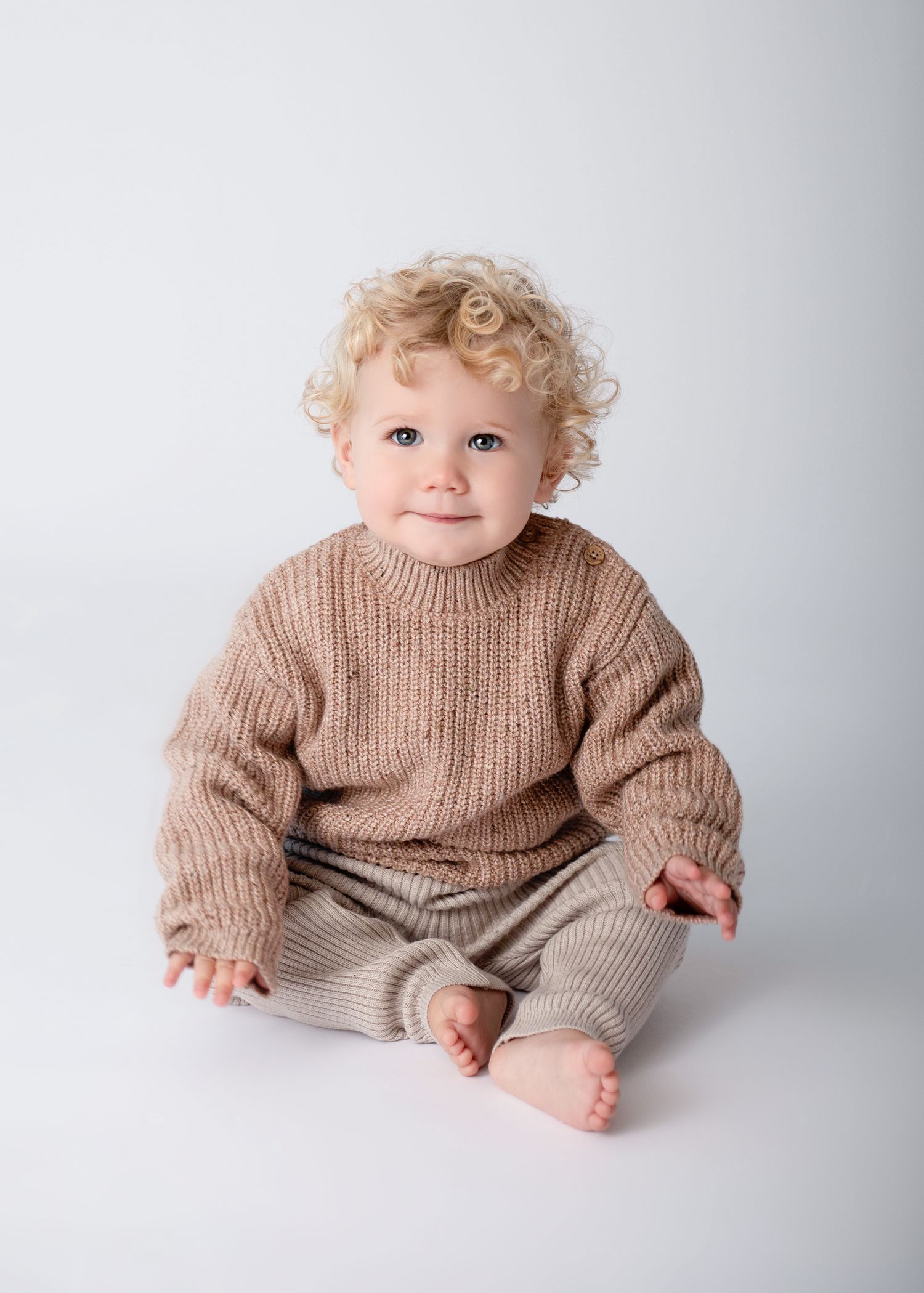 Child in cozy brown sweater and beige pants smiles while sitting on white background.