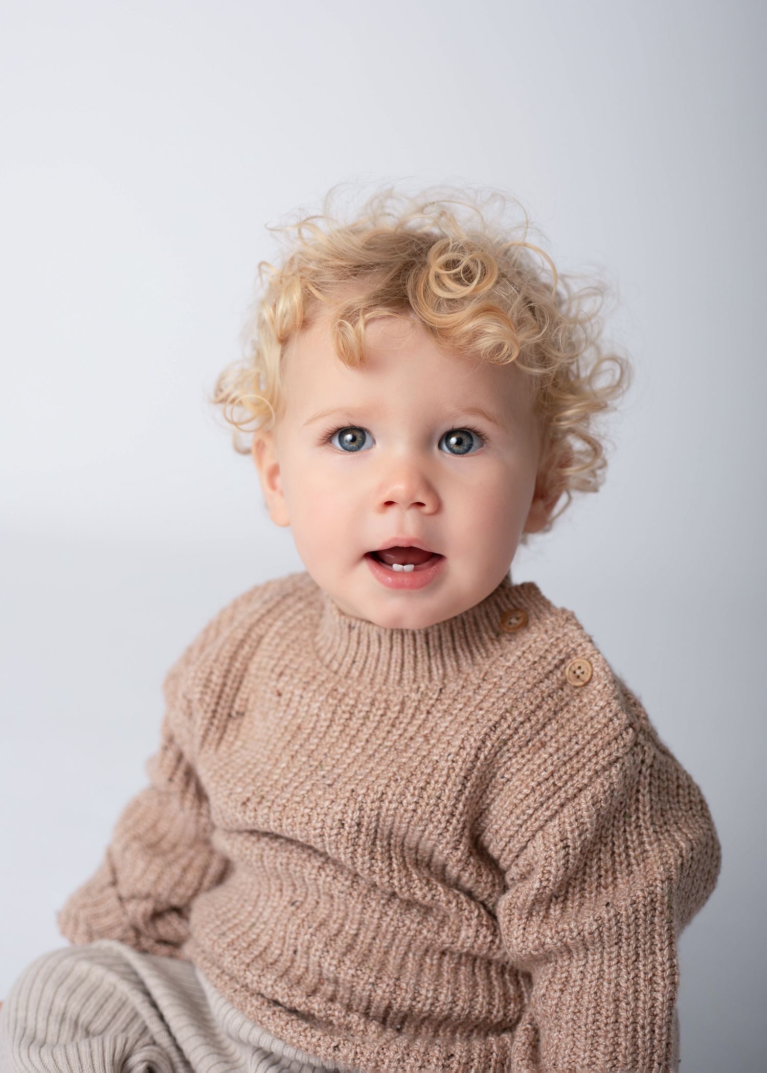 Child in brown knit sweater poses against white backdrop with bright expression.