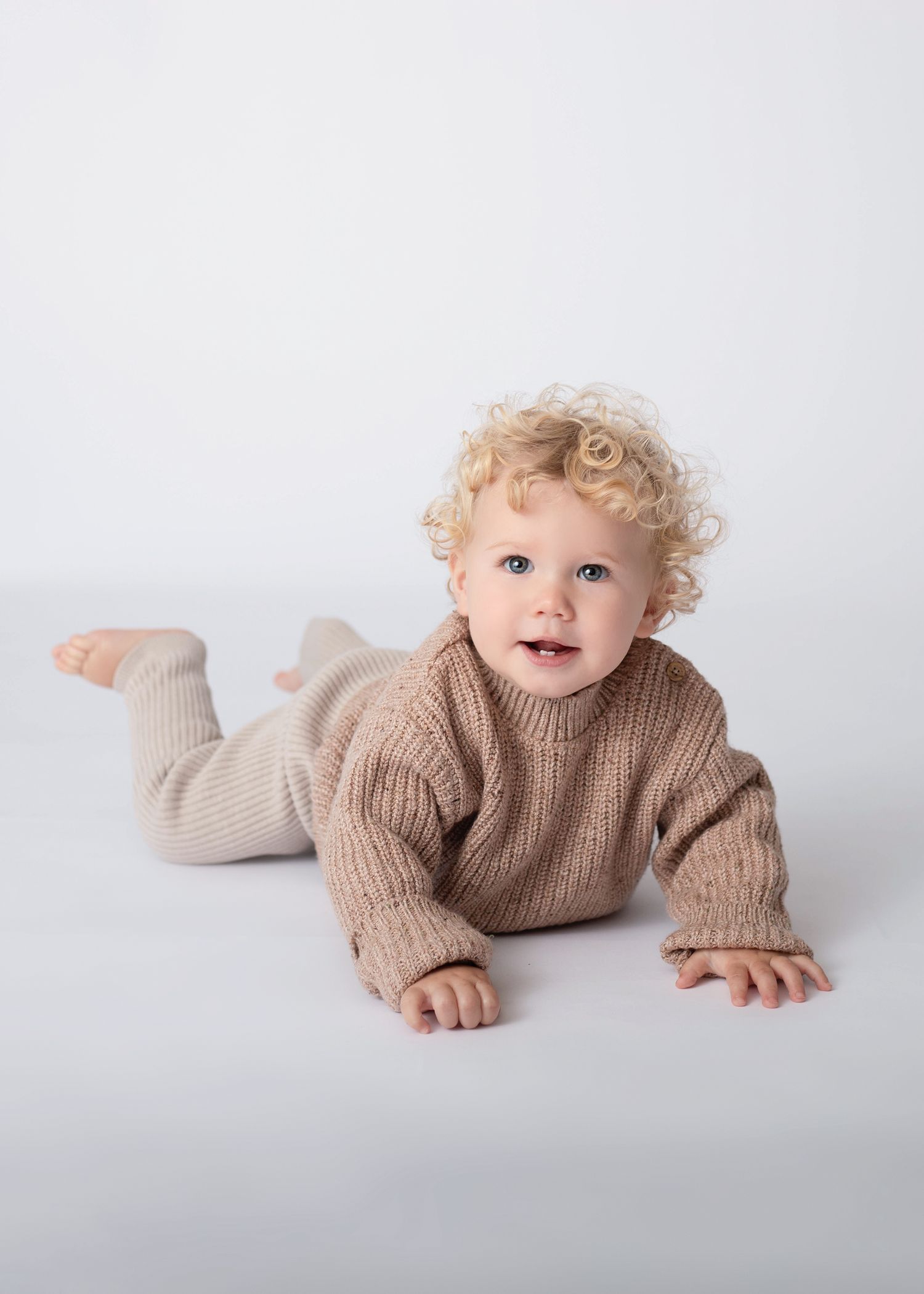 Child in brown sweater lying on stomach on white background with cheerful expression.