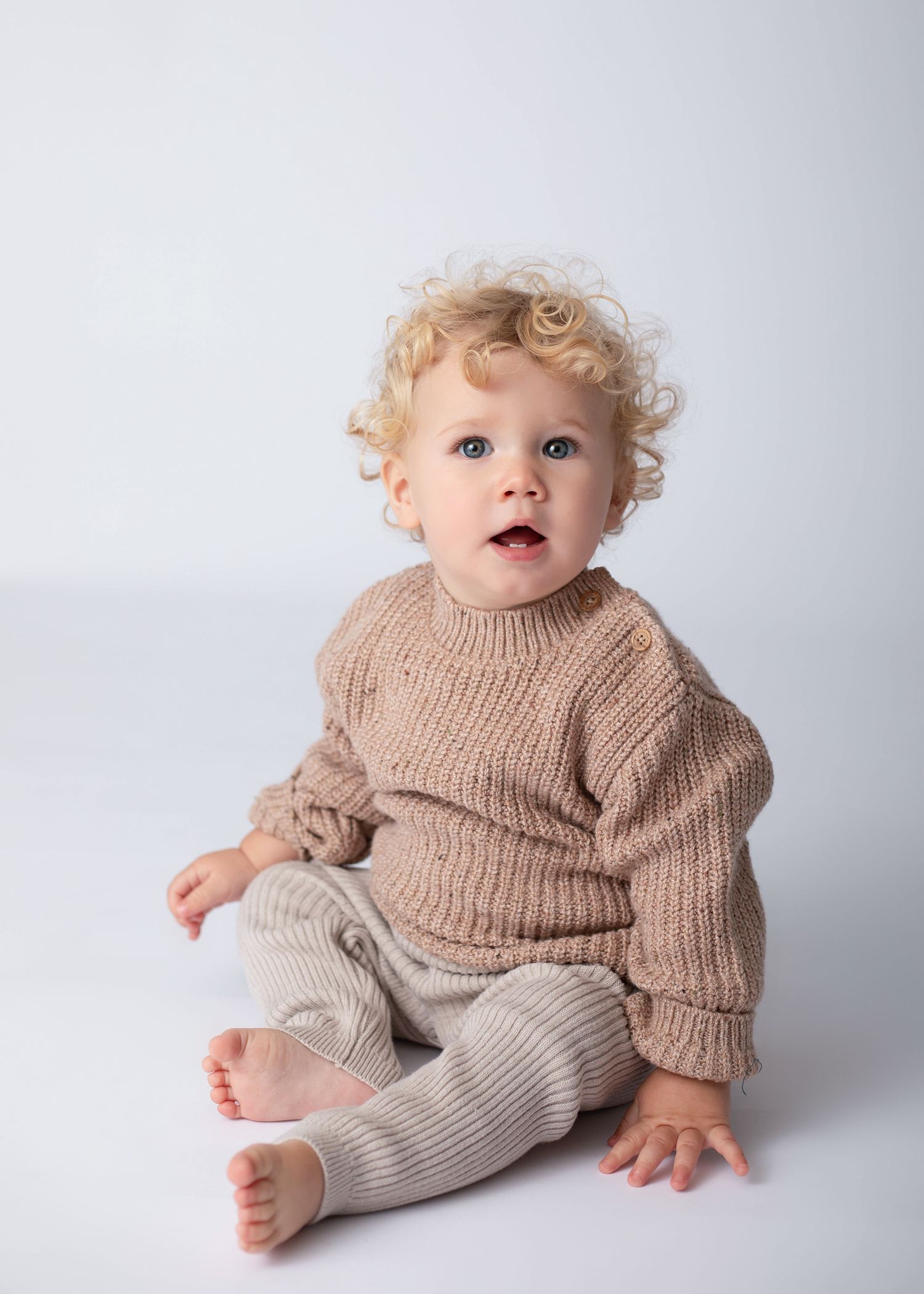 Child wearing brown knit sweater and beige pants sits on white background.