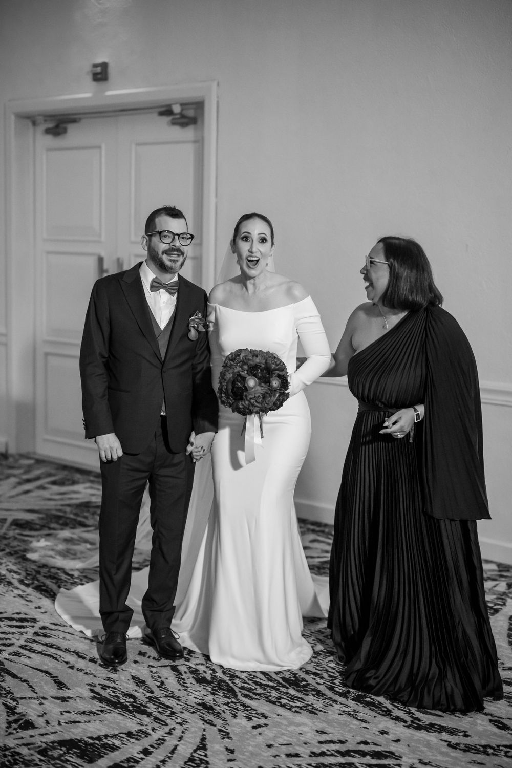 Black and white wedding photo showing three people in formal attire against a patterned carpet.