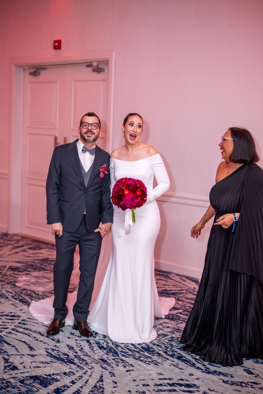 Wedding party members laughing together in a candid moment against pink-lit walls.