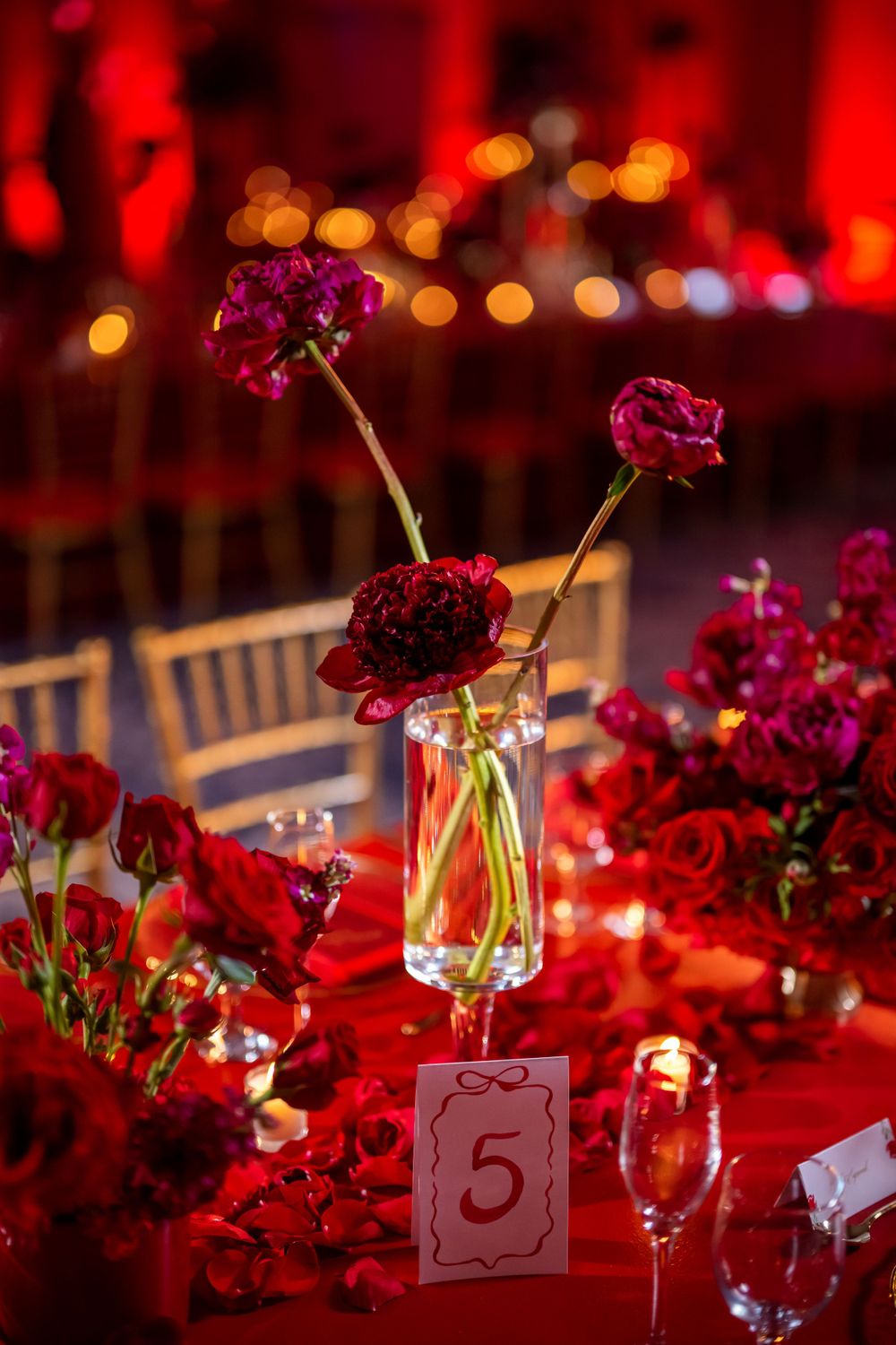 Romantic table setting with red roses in glass vase and table number five against bokeh background.