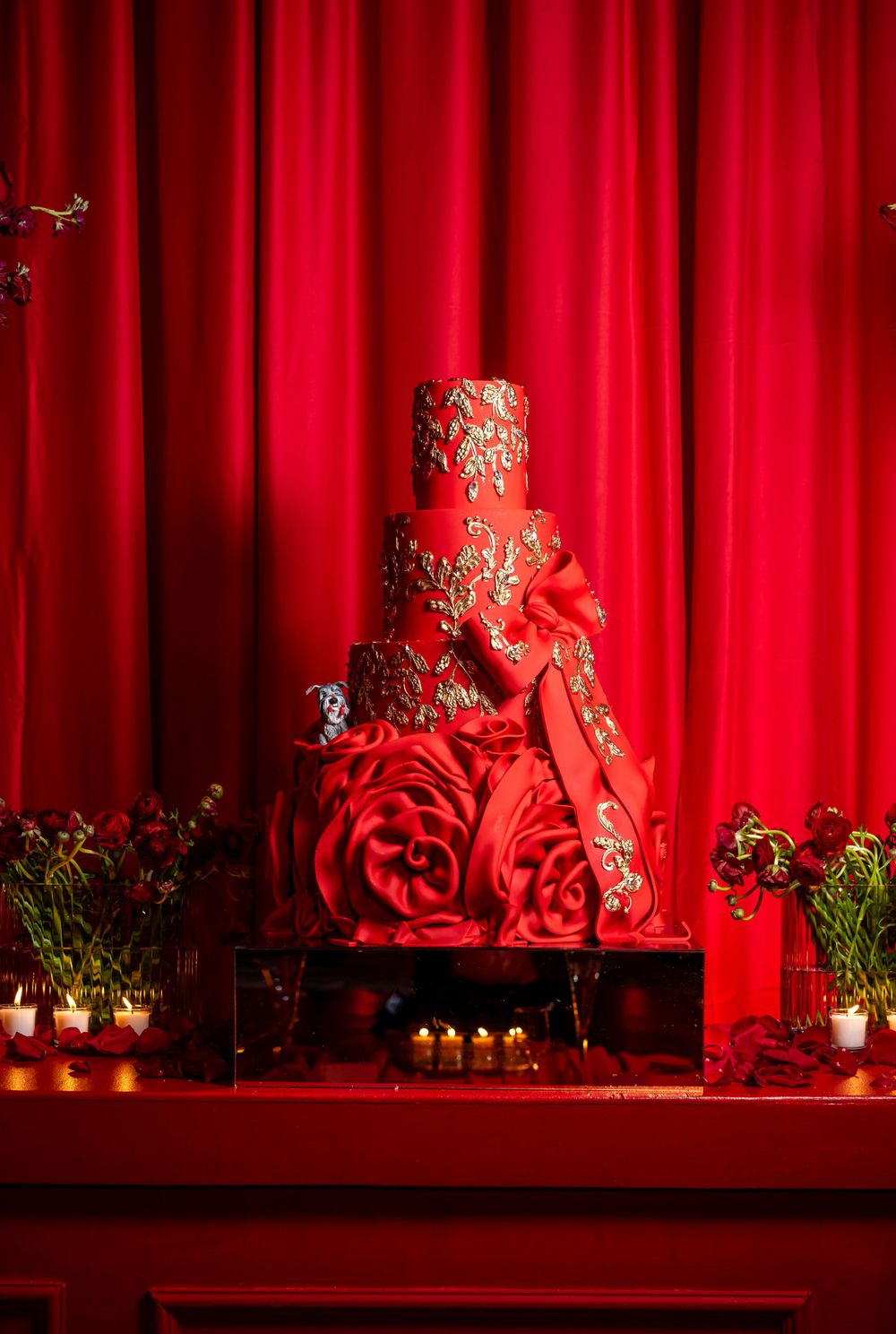 Elegant red wedding cake with gold details and roses displayed against a crimson curtain backdrop.