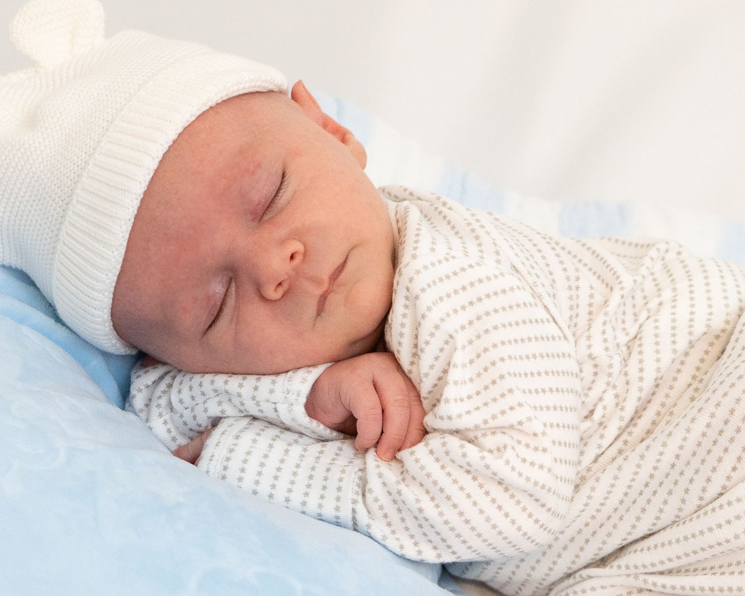 A newborn baby wearing a white knitted outfit sleeps peacefully on blue bedding.