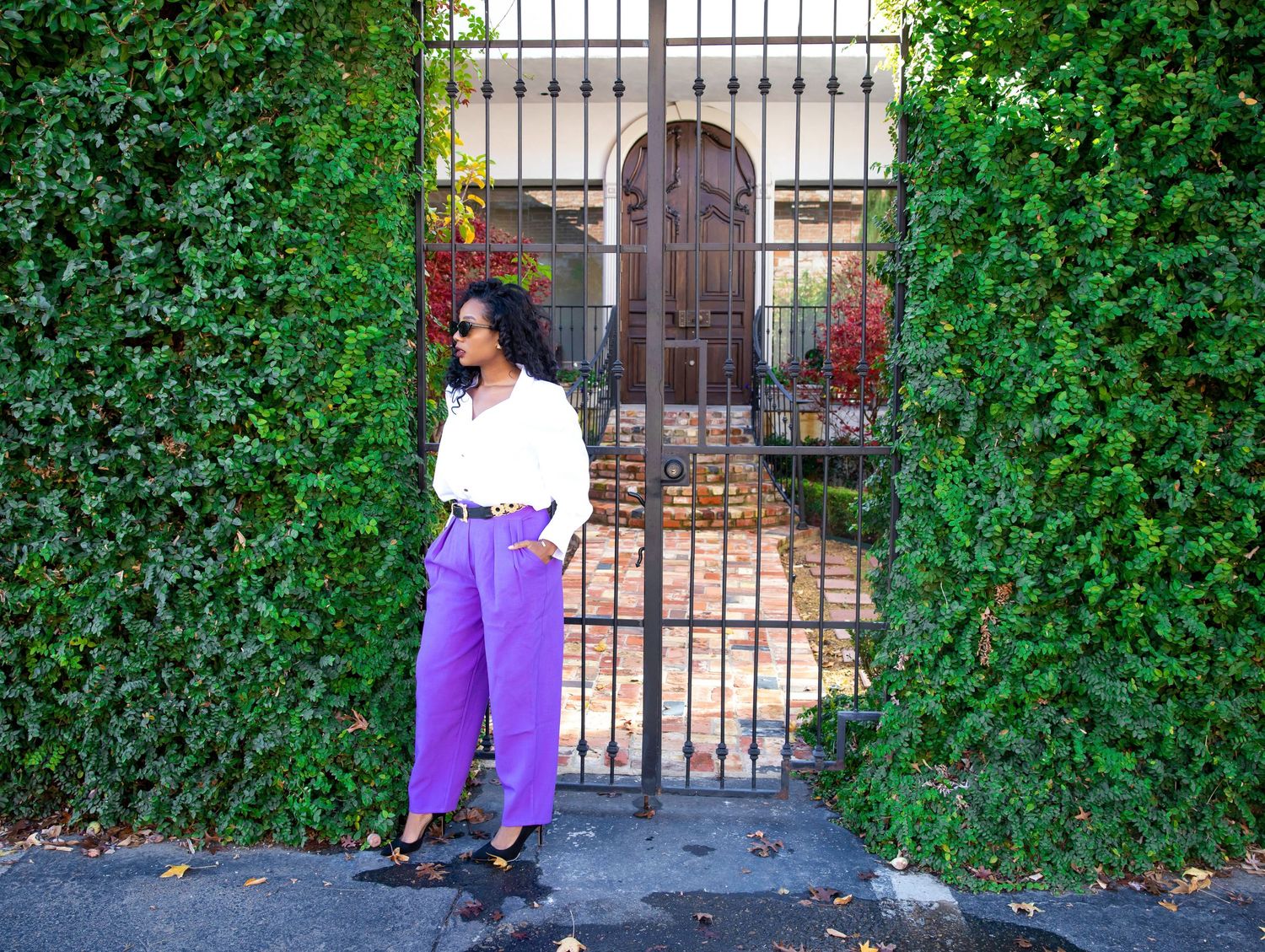 Someone poses by a garden gate with tall hedges wearing a white blouse and purple pants on a sunny day.