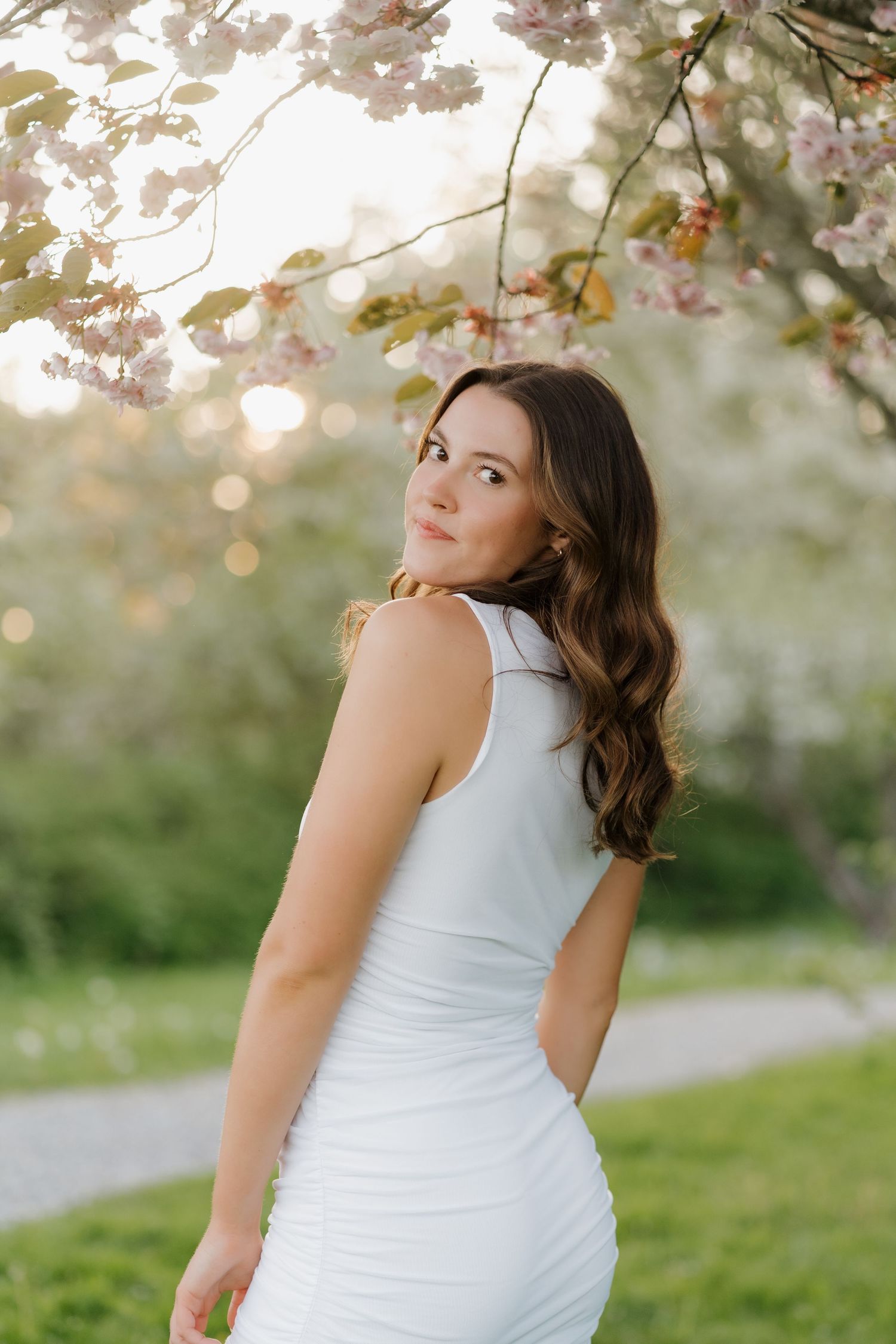 Figure in white dress poses beneath blooming cherry blossoms on a spring day in the park.