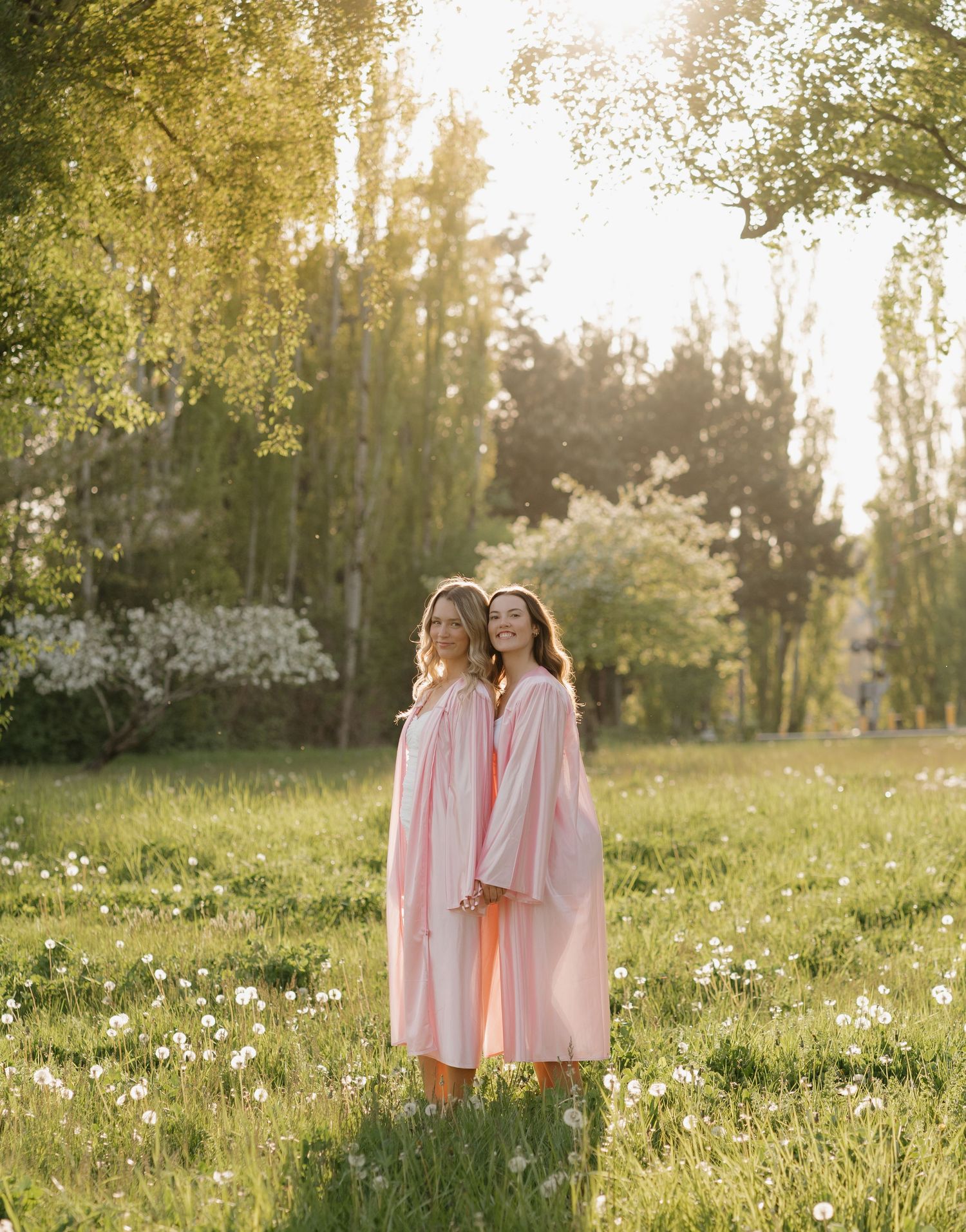 Two people in pink dresses stand together in a meadow filled with wildflowers during golden hour.