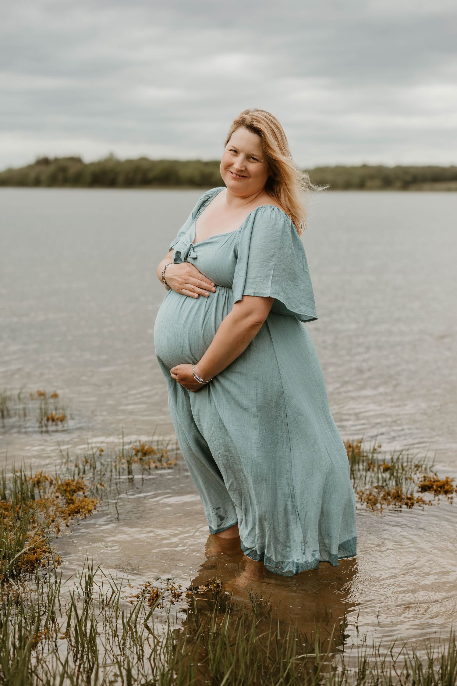 An expectant mother in a teal dress stands in shallow water at sunset.