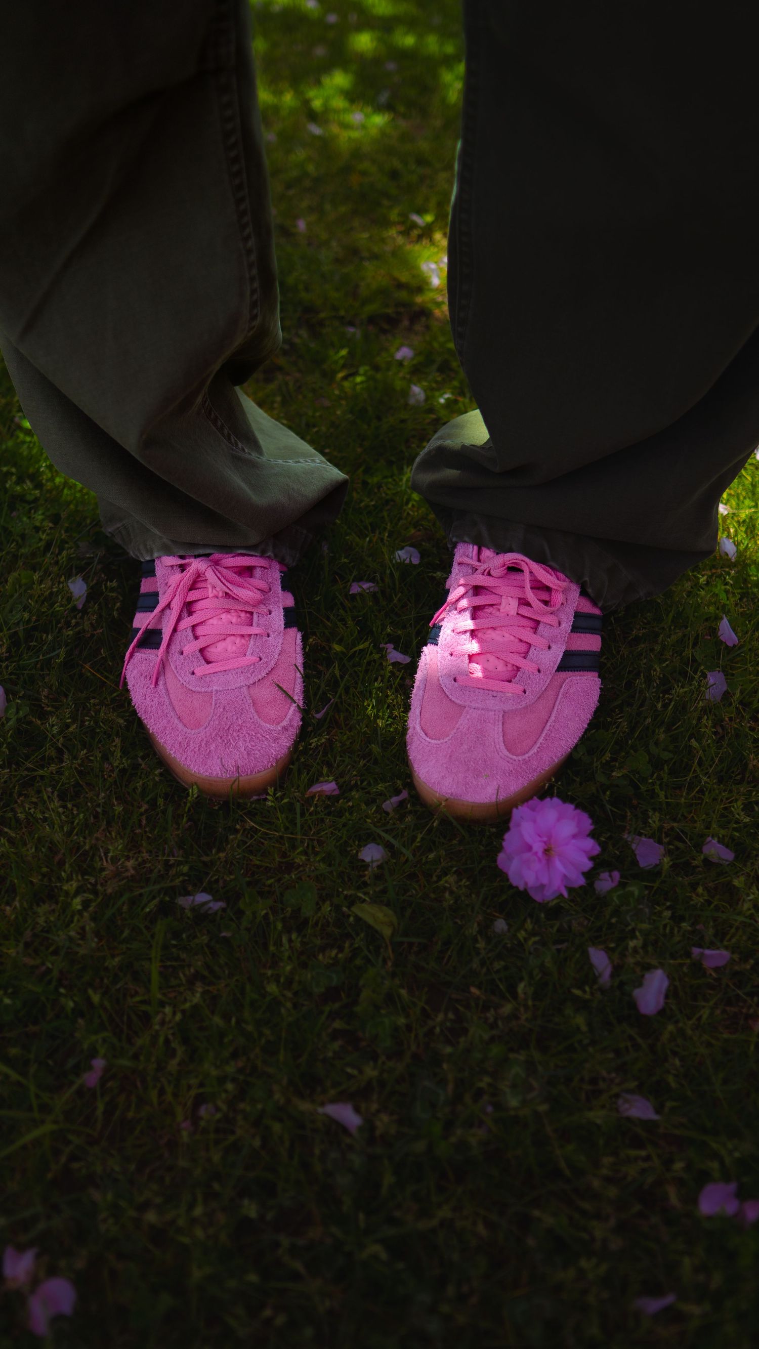 Pink sneakers on grass with fallen purple flower petals in moody lighting.