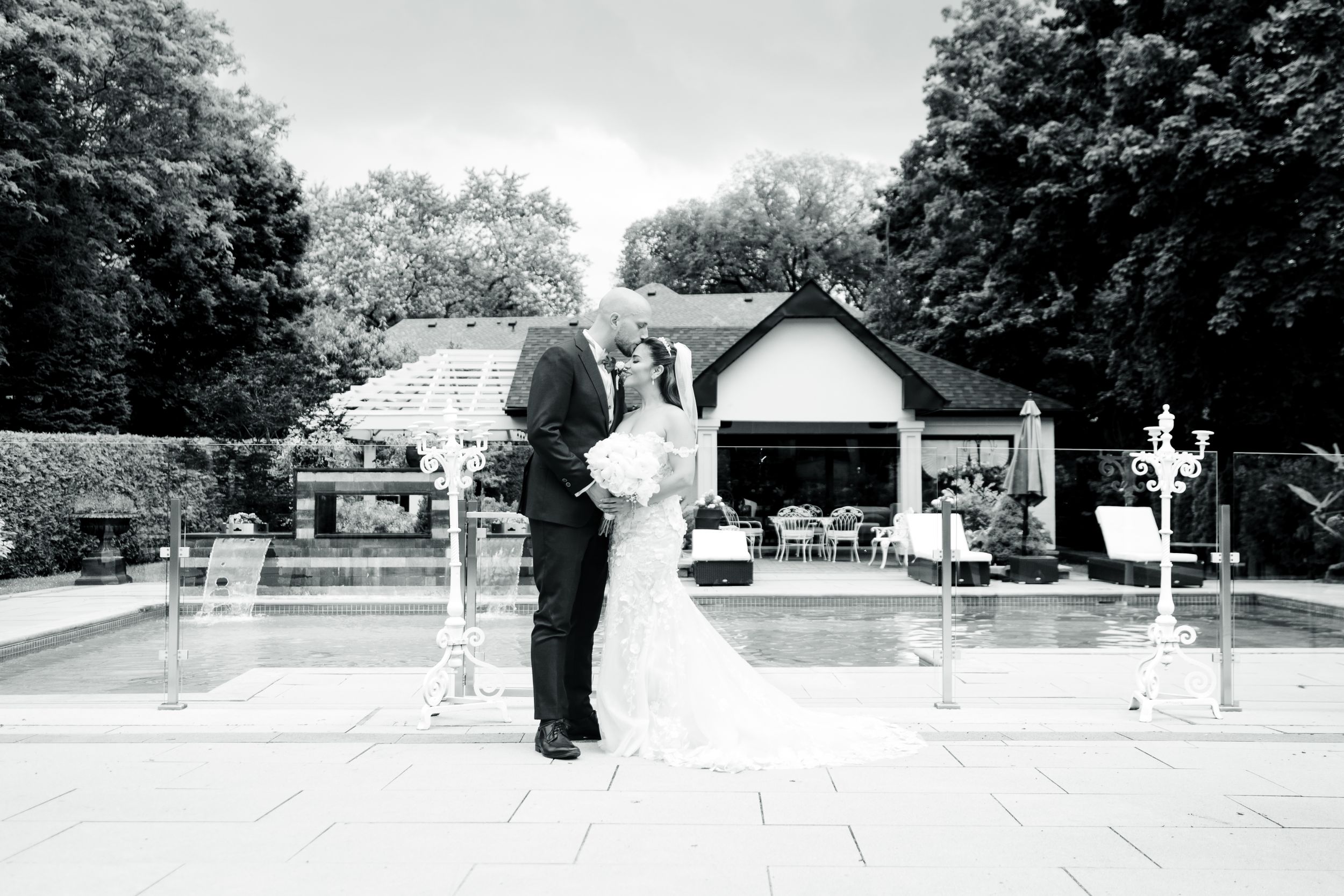Wedding couple stands together outside a white chapel building with trees in background.