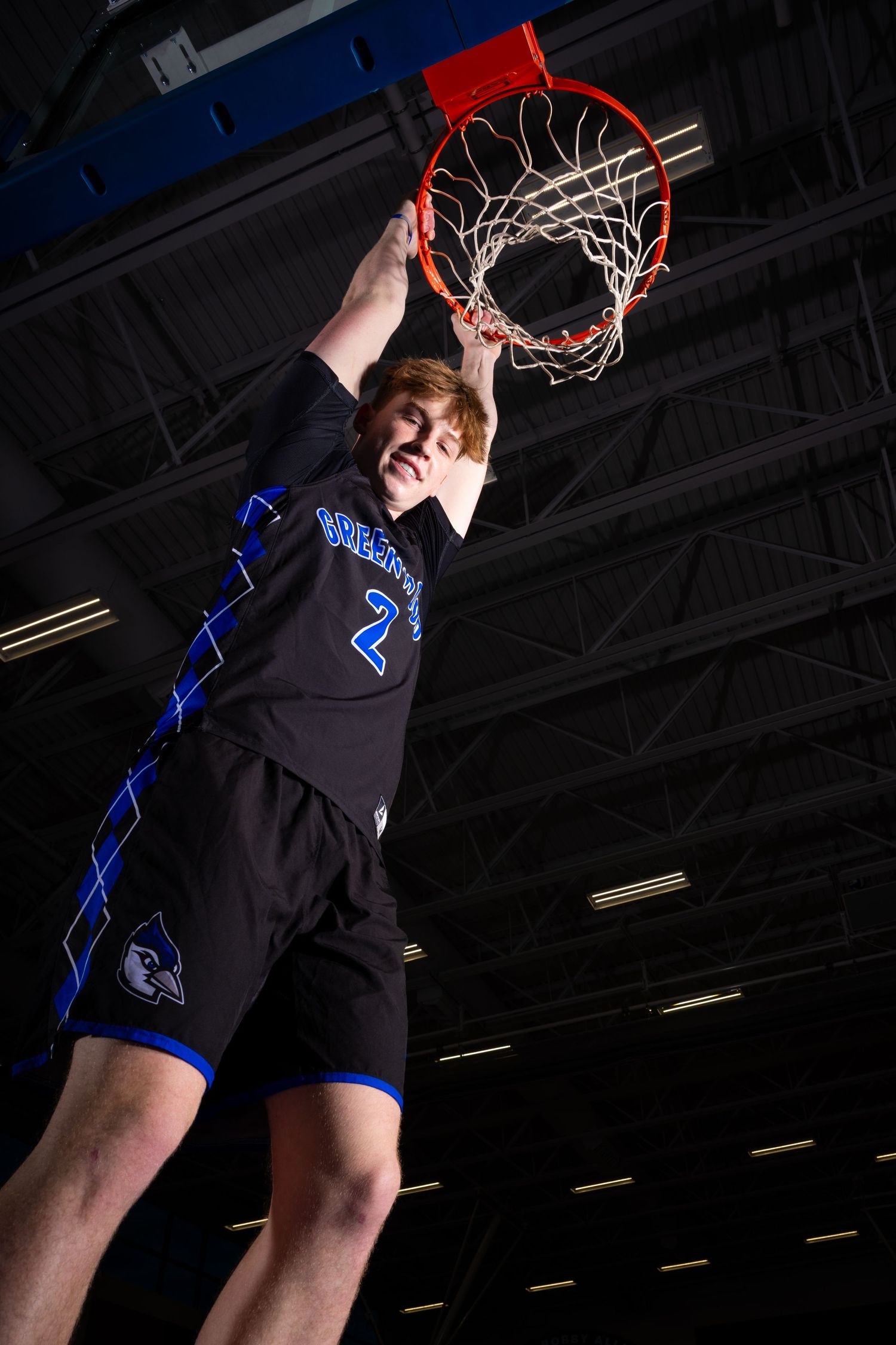 Basketball player jumping high to dunk at the net in a dark indoor gymnasium.